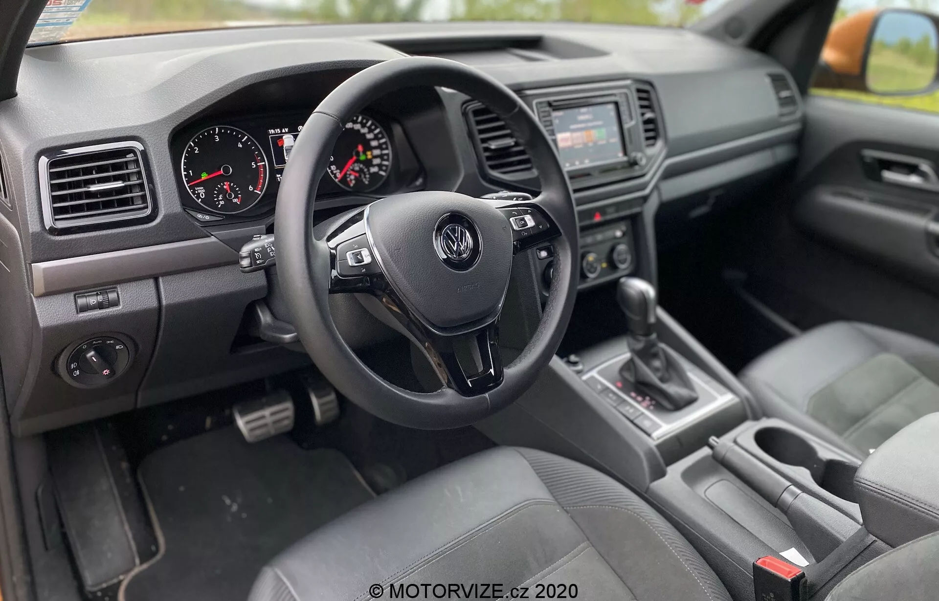 Vista interior del VW Nutzfahrzeuge Amarok 2016 desde el lado del pasajero, mostrando un diseño de conducción a la derecha con un volante Volkswagen de tres radios con botones de control, un cuadro de instrumentos con velocímetro y tacómetro analógicos, una consola central con sistema de audio y pantalla de infoentretenimiento, palanca de cambios en posición 'P', asientos tapizados en tela, un tablero con salidas de aire y plásticos texturizados, y un panel en la puerta del conductor con controles de ventanas eléctricas y ajuste de espejos.