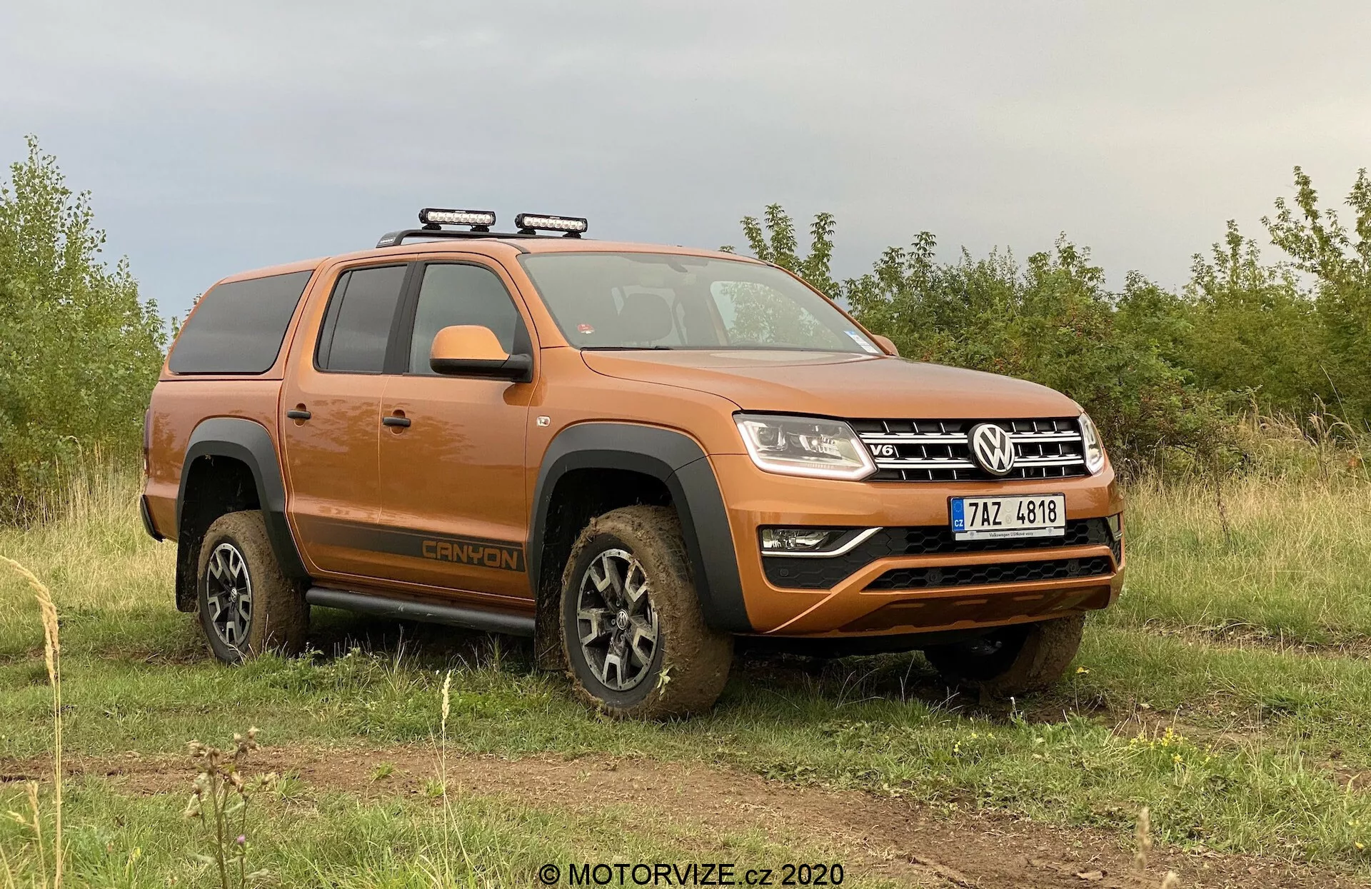 La imagen presenta una camioneta Volkswagen Amarok 2016 de color naranja aparcada en un terreno irregular con pasto y un cielo nublado de fondo. La perspectiva lateral-frontal se enfoca en el lado del conductor, mostrando el perfil del vehículo y parte del frente. Las características clave visibles incluyen un portaequipajes, ruedas cubiertas de barro que indican actividad todoterreno reciente, estribos laterales, configuración de doble cabina, diseño de faros modernos, y un parachoques delantero con faros antiniebla y una placa protectora inferior. El vehículo emana una estética utilitaria todoterreno con elementos tecnológicos modernos.