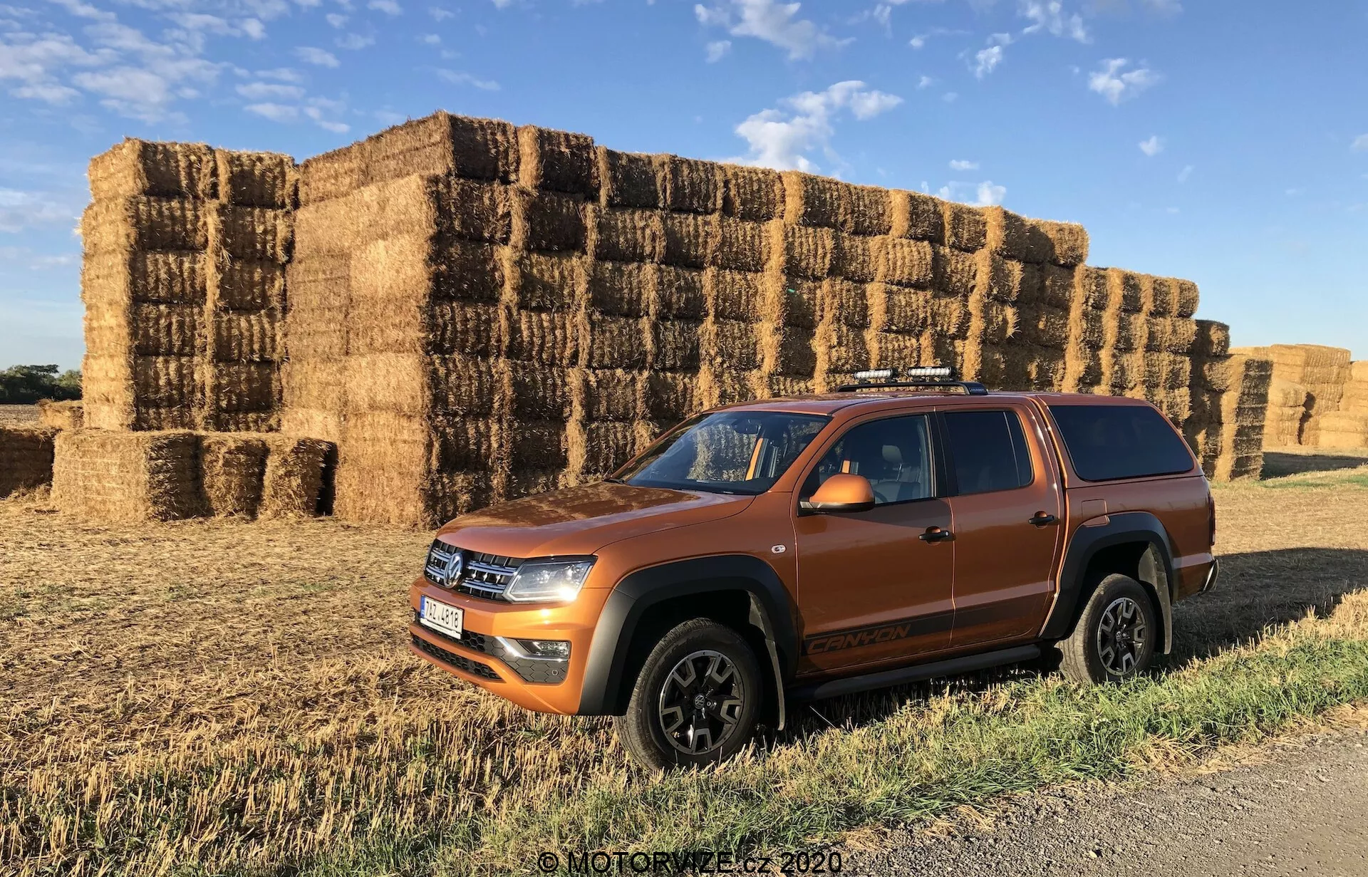 Vista de perfil lateral de un SUV VW Nutzfahrzeuge Amarok 2016 naranja, destacando la fascia frontal, el lado del conductor y el techo. El cuerpo aerodinámico, los faros LED, los arcos de rueda robustos y las ruedas con acabado negro sugieren un diseño moderno y deportivo. Los rieles del techo y la altura libre del suelo visible enfatizan su capacidad multipropósito y todoterreno. Posicionado en un camino de tierra con fardos de heno al fondo, la imagen realza el entorno rústico y robusto del vehículo.