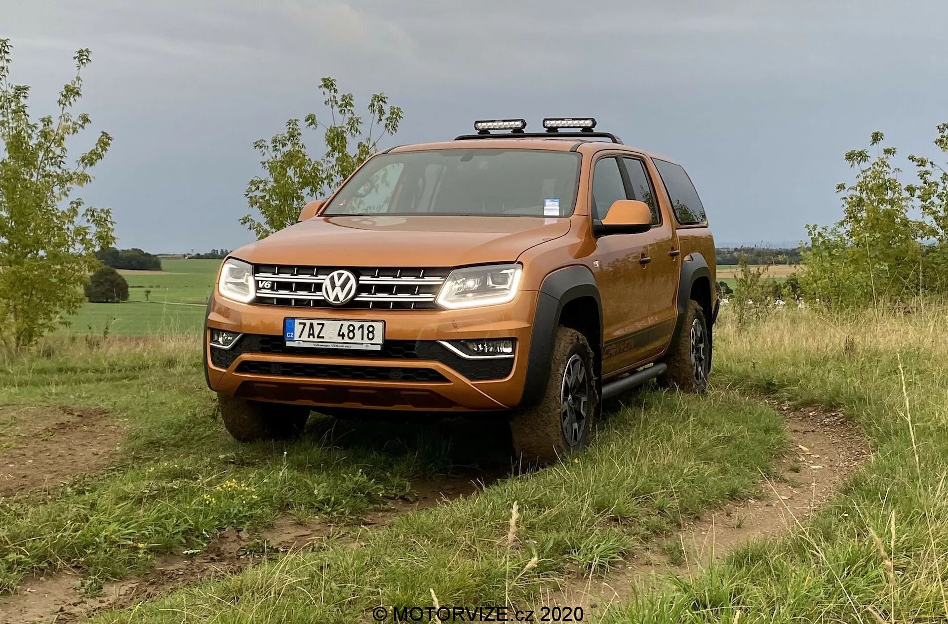 Un SUV Volkswagen naranja, específicamente el VW Amarok 2016, se muestra en un terreno todoterreno cubierto de hierba. La toma desde un ángulo bajo a medio frontal enfatiza su diseño robusto, adecuado para actividades todoterreno. El vehículo presenta una parrilla frontal audaz con dos barras cromadas enmarcando el logotipo central de Volkswagen, un parachoques considerable con una posible placa de deslizamiento, faros compuestos claros con posibles bombillas LED o halógenas y arcos de rueda ensanchados que albergan neumáticos todoterreno. Las luces auxiliares están montadas en el techo, lo que sugiere una funcionalidad adicional para la conducción nocturna. Los detalles visibles incluyen el parabrisas, los rieles de techo, los espejos laterales con indicadores de señal integrados y la matrícula delantera. El vehículo parece limpio con algo de suciedad en los lados y ruedas inferiores, lo que encaja con el uso todoterreno. El fondo sugiere un entorno rural o semi-rural.