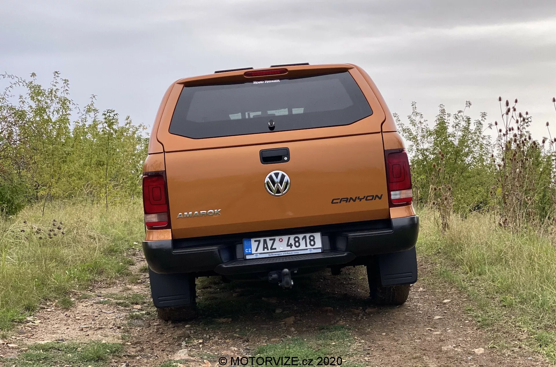 Vista trasera profesional de una camioneta Volkswagen Amarok 2016 desde un ángulo bajo, destacando la robusta puerta trasera con la marca 'AMAROK', el logo de Volkswagen centrado, indicador de nivel de acabado 'CANYON', placa de matrícula, luces traseras detalladas, parachoques con sensores de estacionamiento y escalón, barra de remolque, estacionada en un camino sin pavimentar que indica capacidad todoterreno.