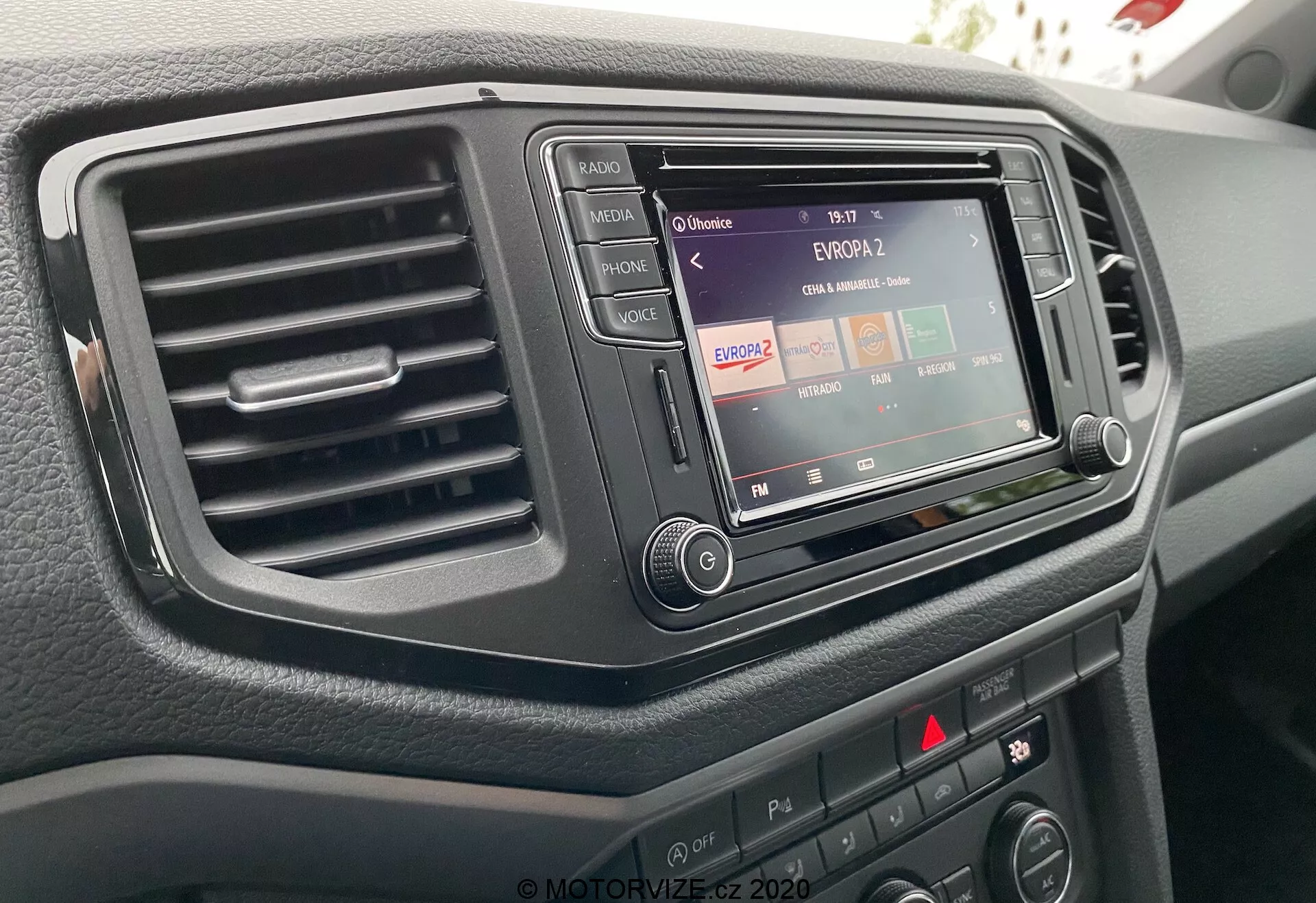 Imagen en primer plano de la consola central de un VW Nutzfahrzeuge Amarok 2016 desde la perspectiva del conductor, destacando el sistema de infoentretenimiento con funcionalidad de radio, una sola ventilación a la izquierda y varios botones de control y perillas. El sistema de infoentretenimiento está incrustado en un ribete oscuro y elegante, y la superficie del salpicadero tiene un acabado texturizado.