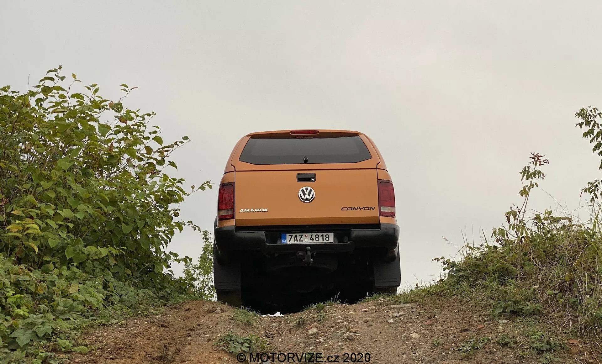 Vista trasera de un crossover o SUV Volkswagen naranja desde un ángulo ligeramente bajo, capturada en la cresta de una pequeña colina. Los elementos de diseño trasero del vehículo, como la puerta trasera, la ventana trasera, las luces traseras, las insignias de marca y modelo que leen 'Volkswagen' y 'CROSSOVER,' y el parachoques trasero son claramente visibles. La matrícula es visible, pero no se proporcionan detalles específicos. La puerta trasera tiene un pliegue prominente y líneas de diseño, con un posible asa o hueco debajo de la matrícula. Las luces traseras envuelven hacia los lados e incluyen indicadores, luces de reversa y luces de freno. La parte inferior negra del parachoques trasero sugiere un revestimiento protector para uso todoterreno. El vehículo está enmarcado por vegetación, lo que indica un entorno rural o todoterreno. El suelo es de tierra natural, consistente con las capacidades todoterreno del vehículo. En la esquina inferior derecha hay una marca de agua que dice 'MOTORVIZE.cz ©2020'.