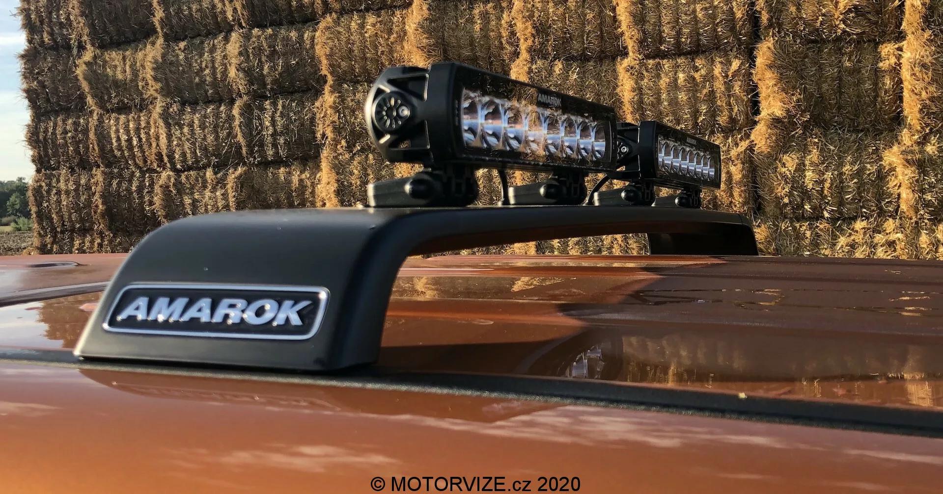 Vista en primer plano de una camioneta Volkswagen Amarok 2016 que muestra una barra de luz LED aftermarket montada en el techo. La carcasa negra de la barra de luz con elementos LED individuales es claramente visible, con la marca 'AMAROK' en una cubierta ventilada. La foto, tomada al aire libre, muestra las lentes reflectantes de la barra de luz y pacas de heno en el fondo, indicando un entorno rural.