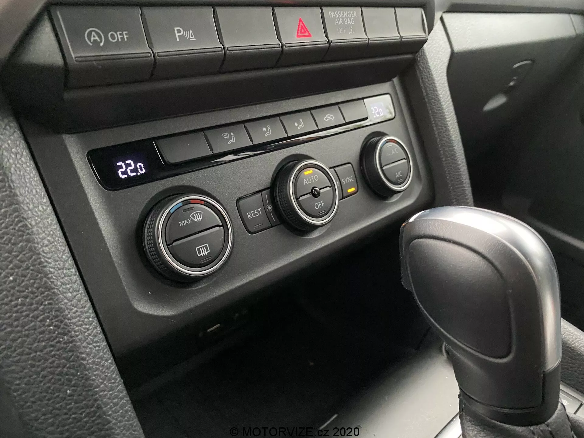 Vista de cerca del sistema de control climático y parte de la palanca de cambios del VW Nutzfahrzeuge Amarok 2016. Perspectiva del pasajero o conductor mirando hacia la consola central. Cuenta con una palanca de cambios automática con textura de cuero en 'Park' y un panel de control climático configurado a 22°C con tres perillas giratorias y varios botones funcionales. Los botones incluyen desactivación de Auto Stop/Start, alternador de sensores de estacionamiento, volante calefactado y asiento calefactado, junto con un botón de luz de emergencia rojo central. La iluminación suave y difusa sugiere un interior bien sombreado.