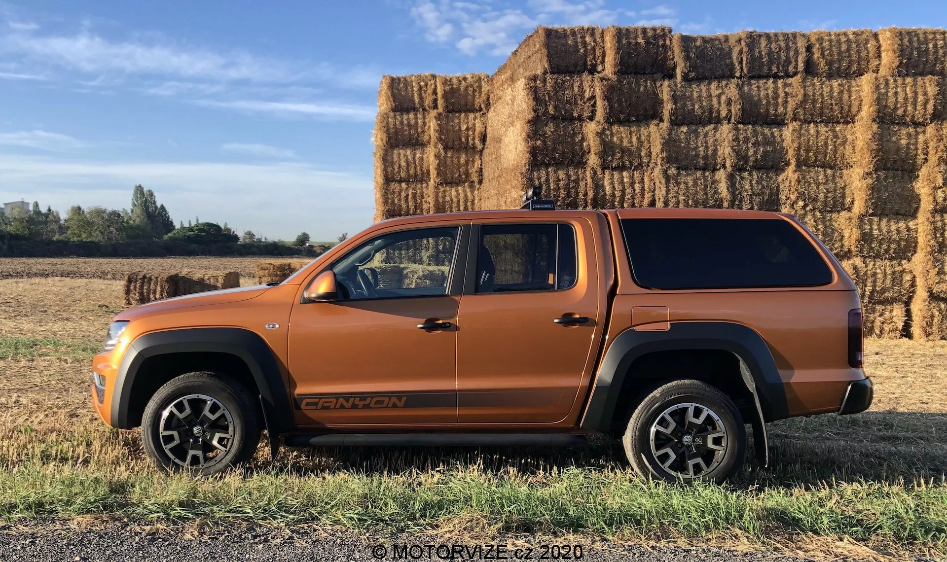 Vista de perfil lateral de un vehículo tipo SUV de color naranja metálico, probablemente un SUV mediano o pickup con cabina extendida y cubierta de la caja del camión, estacionado sobre terreno herboso plano con grandes fardos de heno rectangulares en el fondo bajo un cielo soleado y despejado. El vehículo muestra líneas de carrocería audaces, grandes arcos de rueda, molduras inferiores negras, ruedas robustas de aleación, una insignia visible en la parte baja de la puerta delantera y una antena en el techo. La foto destaca las posibles capacidades todoterreno y el diseño sin costuras de la cubierta de la caja del camión.