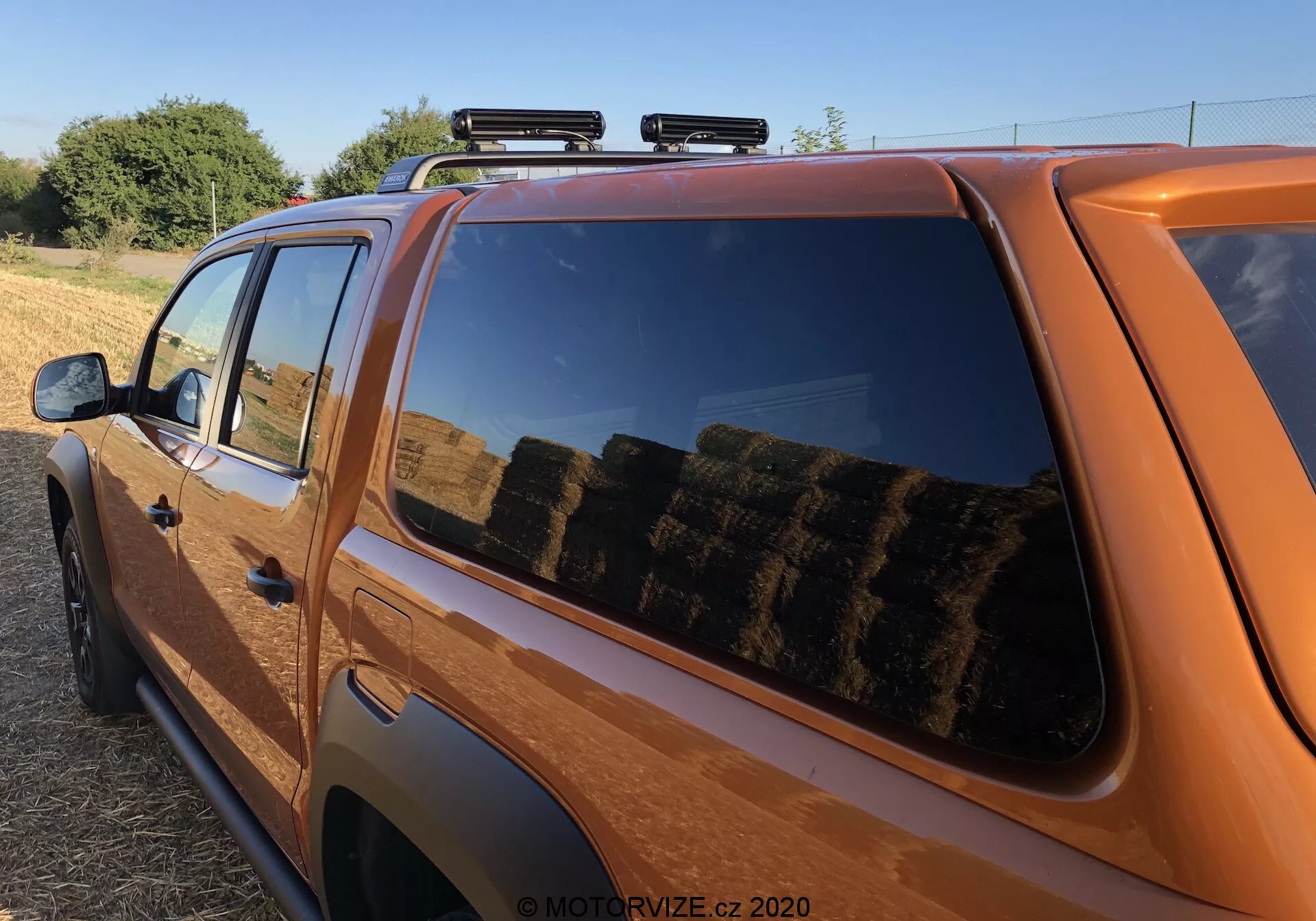 Toma cercana y en ángulo lateral de una camioneta naranja, mostrando predominantemente la sección trasera superior de la cabina y una porción de la cama. Las características incluyen un perfil lateral claro con un enfoque en la ventana trasera del pasajero, reflejando un entorno rural con pacas de heno, portaequipajes para carga extra, diseño moderno y ligeramente tintado de la ventana, pintura brillante, color naranja distintivo, insignia visible, espejo lateral parcial y luz solar cálida que sugiere temprano en la mañana o al atardecer.