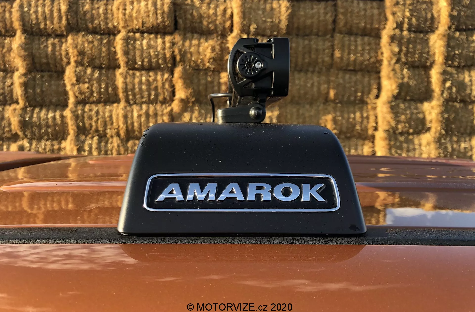 Foto de primer plano de un pickup Volkswagen Amarok 2016 que presenta una cámara de marcha atrás instalada en la luz de freno montada en alto con el nombre 'AMAROK' en letras plateadas en relieve sobre una carrocería de color bronce metálico, al aire libre contra un fondo con patrón de ladrillo.