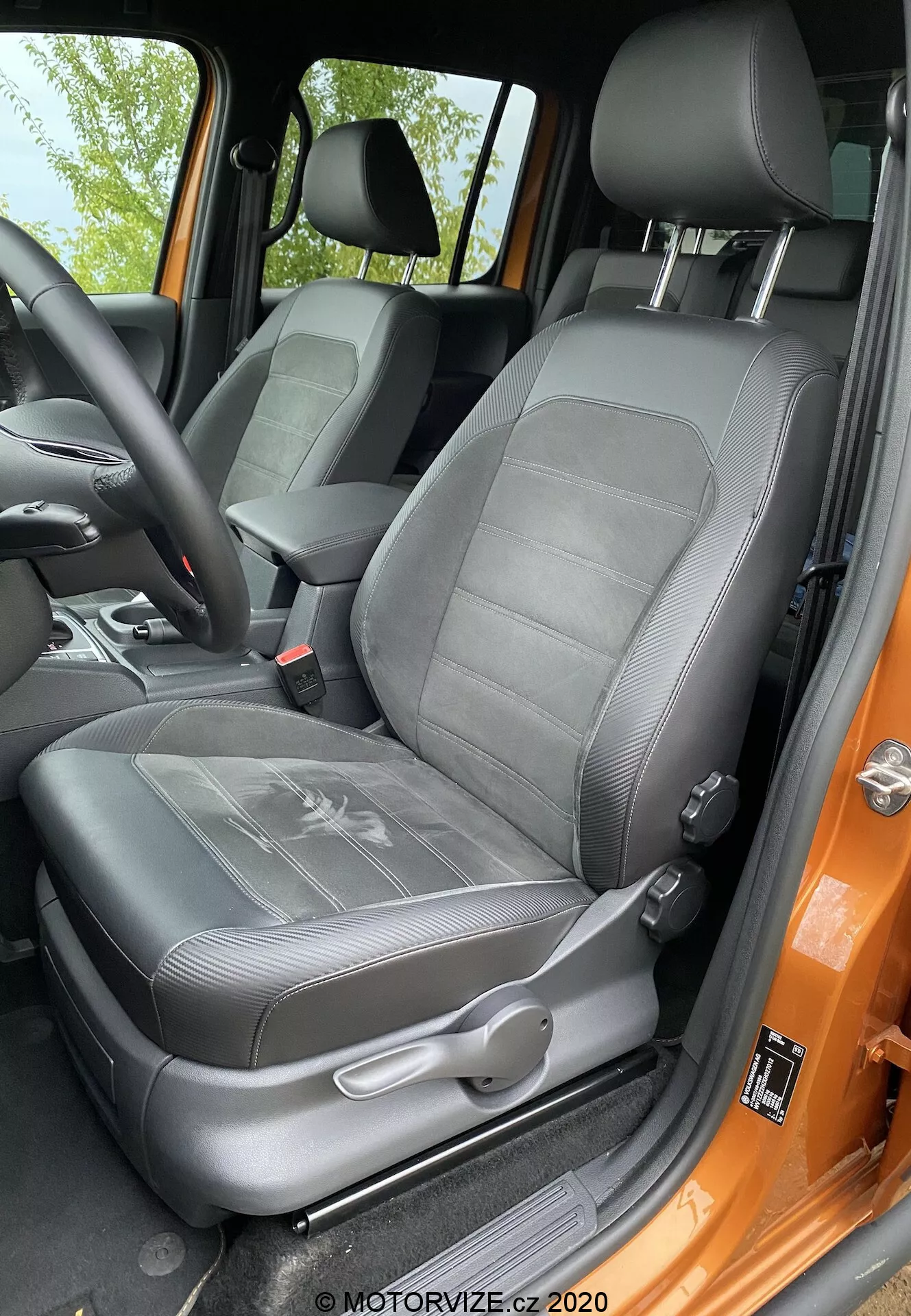 Vista en primer plano del asiento del conductor del VW-nutzfahrzeuge Amarok 2016 desde la perspectiva de alguien parado fuera de la puerta abierta. El asiento tapizado en cuero negro muestra un diseño con texturizado acanalado y un reposacabezas ajustable integrado. Controles de ajuste manual y el mecanismo del cinturón de seguridad de tres puntos son visibles. El soporte lateral indica soporte lateral, y el marco de la puerta pintado en un audaz color naranja contrasta con el interior negro. El asiento parece bien mantenido e iluminado por luz natural.