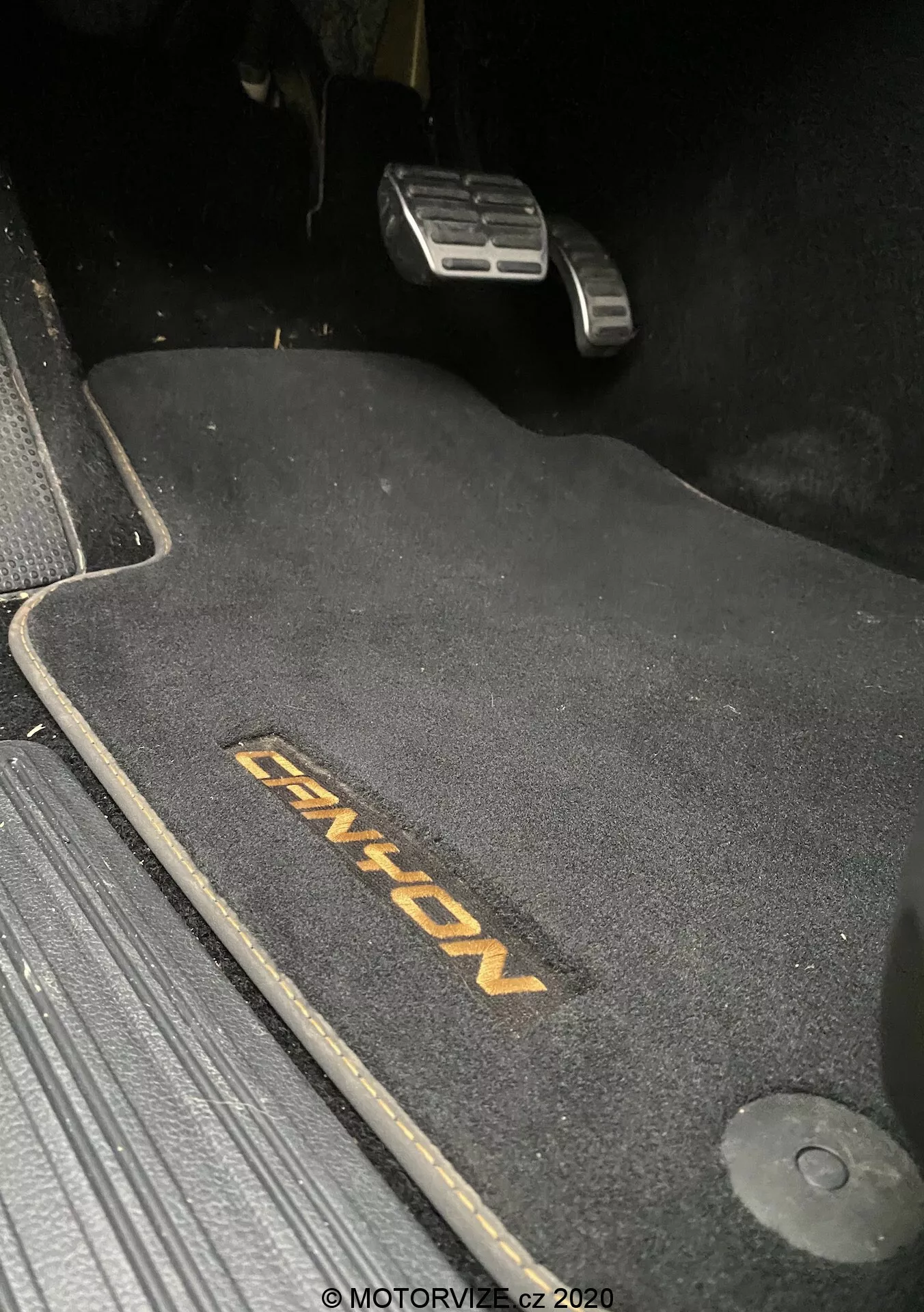 Vista de cerca del área del conductor en el VW-Nutzfahrzeuge Amarok 2016, mostrando una alfombrilla oscura con el bordado 'ENYON'. Son visibles los pedales metálicos del acelerador y del freno, con el pedal de freno más alto, lo que sugiere que está activado. La alfombrilla está diseñada para adaptarse al espacio, con un gancho de retención a la derecha. La alfombrilla contrasta con los alrededores oscuros, indicando un interior de tema oscuro. La escena está ligeramente iluminada, probablemente tomada por la noche o en condiciones de poca luz.