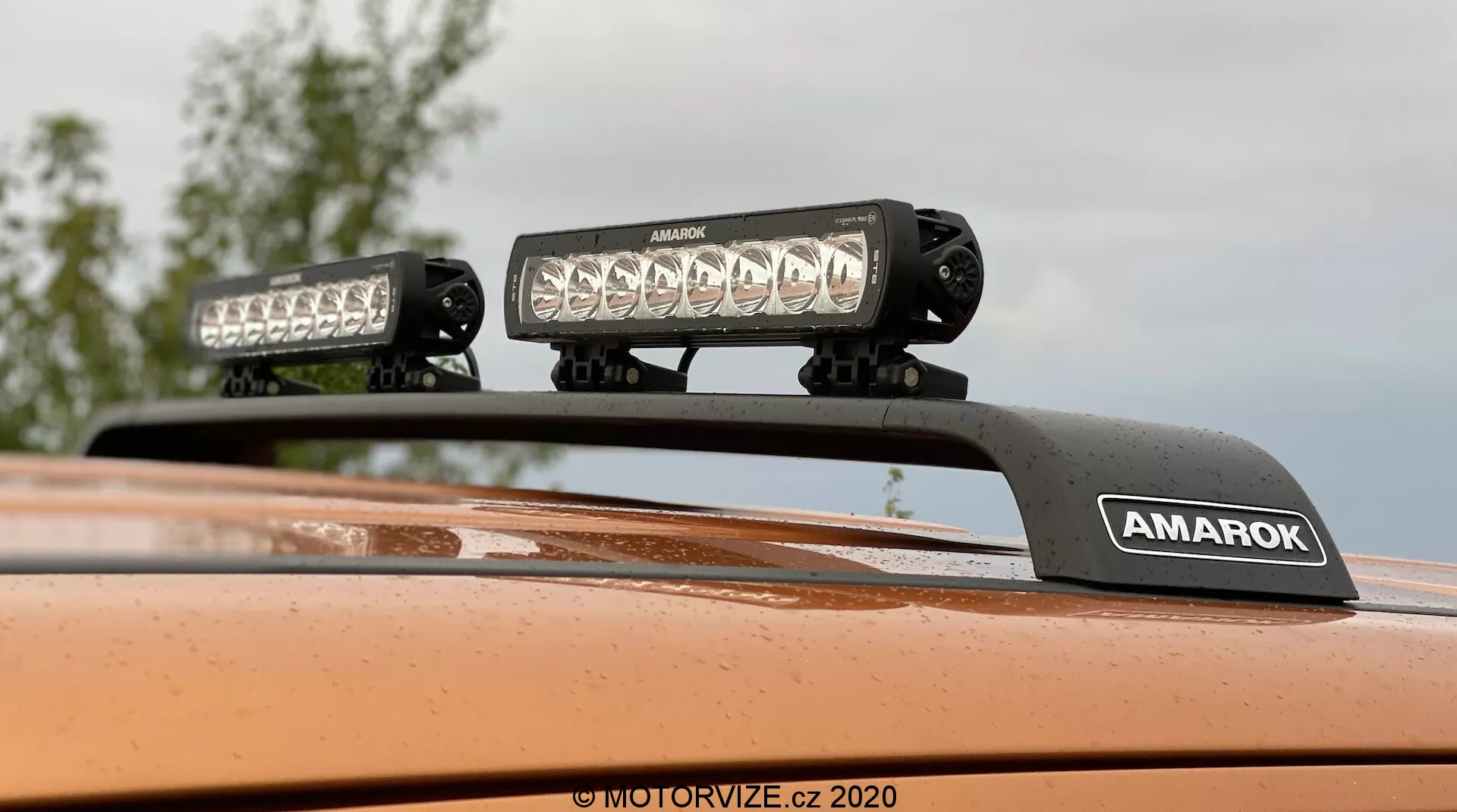 Primer plano enfocado hacia arriba del techo de un Volkswagen Amarok 2016 con equipamiento de iluminación montado en el techo, dos barras de luz LED de alta intensidad en un soporte con la marca 'AMAROK', acabado de pintura metálica naranja; ideal para características de vehículos todoterreno o accesorios para vehículos.