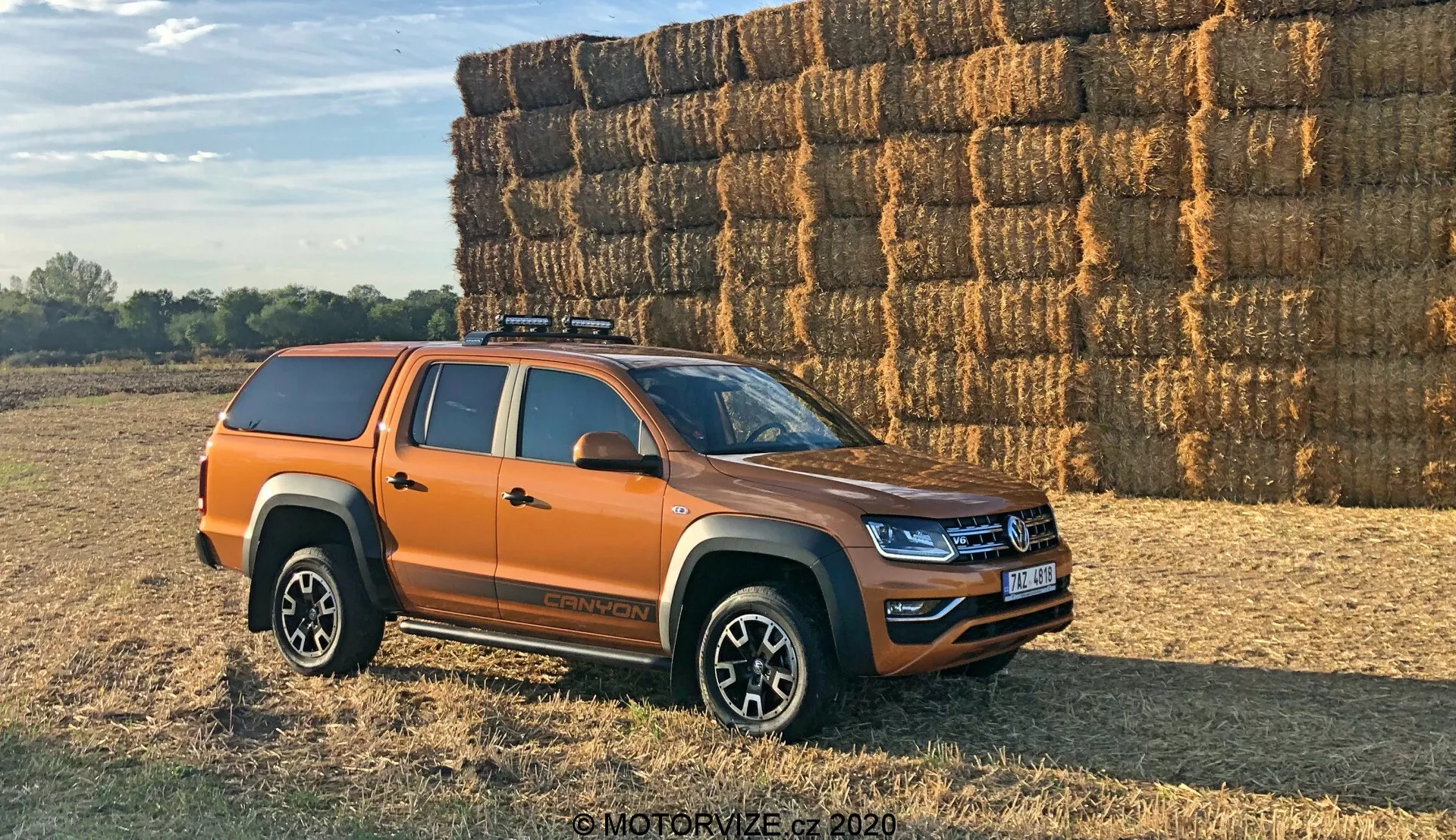 Vista lateral de una camioneta Volkswagen Amarok naranja estacionada al aire libre sobre una superficie desigual, posible de tierra o césped. El vehículo está posicionado a un ángulo de 30 grados hacia la cámara, mostrando el perfil lateral y una vista parcial de la parrilla frontal y los faros. La camioneta moderna de tamaño medio tiene líneas de diseño frescas y iluminación LED, lo que sugiere un modelo reciente. Se ven cuatro puertas que indican una configuración de doble cabina, llantas de aleación multirradio, neumáticos todoterreno, una cubierta dura sobre la caja de la camioneta y un sistema de portaequipajes en el techo. Los espejos laterales probablemente incluyen indicadores. Fotografiado en una luz cálida diurna con sombras largas, un gran montón de pacas de heno en el fondo sugiere un entorno rural.