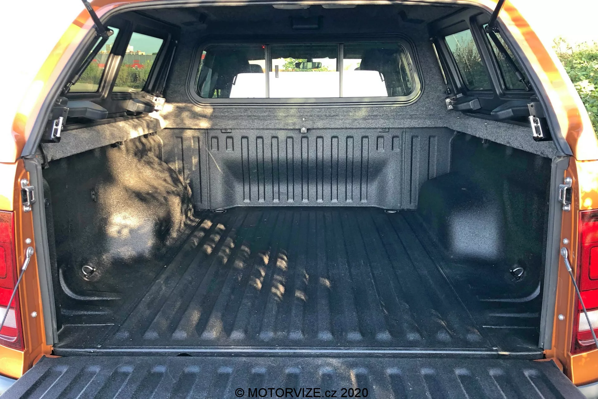 Interior del espacio de carga del VW Nutzfahrzeuge Amarok 2016, visto desde la parte trasera con la puerta trasera abierta, mostrando el área del maletero. El área de carga cuenta con un forro resistente y texturizado, paredes laterales terminadas con material duradero, puntos de amarre para asegurar objetos, iluminación interior para visibilidad, asientos traseros visibles en la parte frontal del espacio y un borde protector en el borde de la puerta trasera.