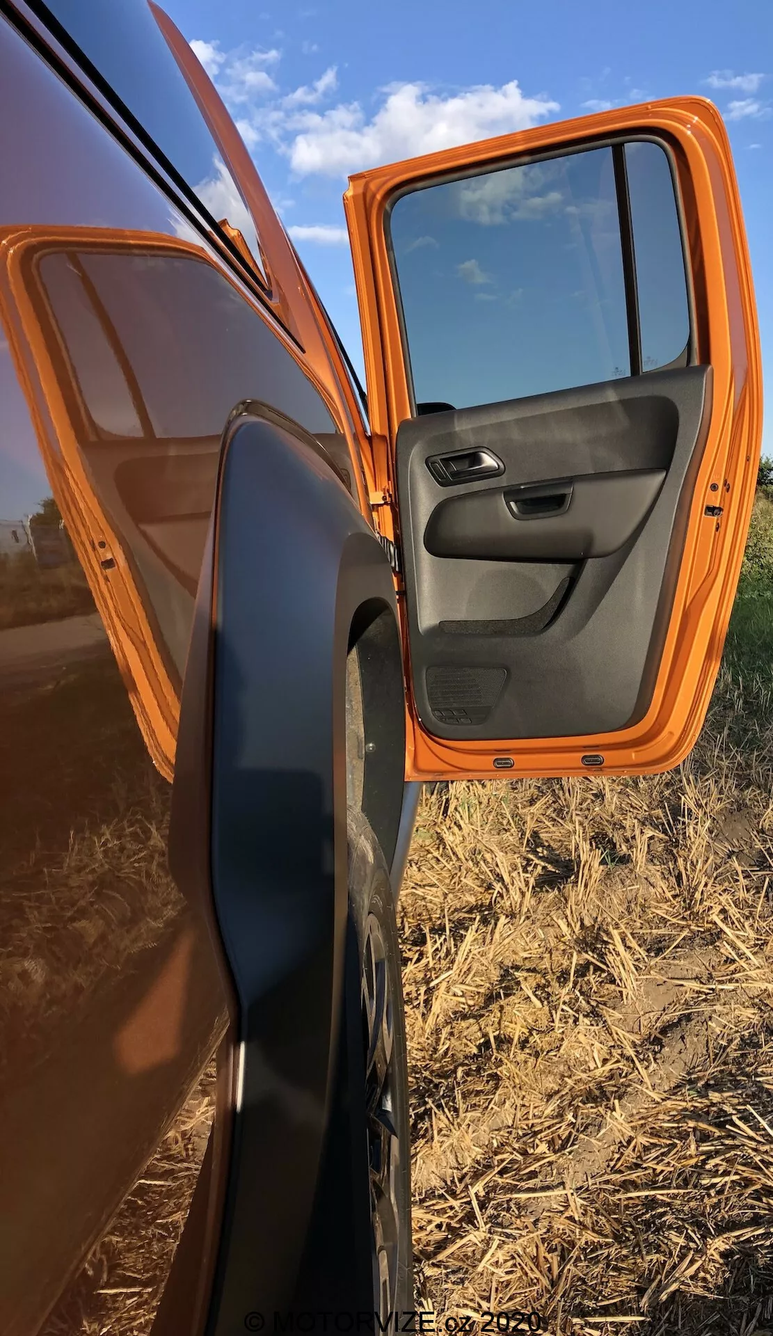 Vista de cerca de una puerta delantera del pasajero parcialmente abierta de un VW Nutzfahrzeuge Amarok 2016 naranja, mostrando la pintura exterior naranja brillante y el panel interior negro con controles integrados. El coche está estacionado en diagonal, presentando un perfil lateral con la puerta abierta en un ángulo de 70 grados, destacando el espejo lateral con carcasa negra. Los reflejos en la puerta muestran un cielo despejado y tierras de cultivo planas, acentuando el entorno rural.