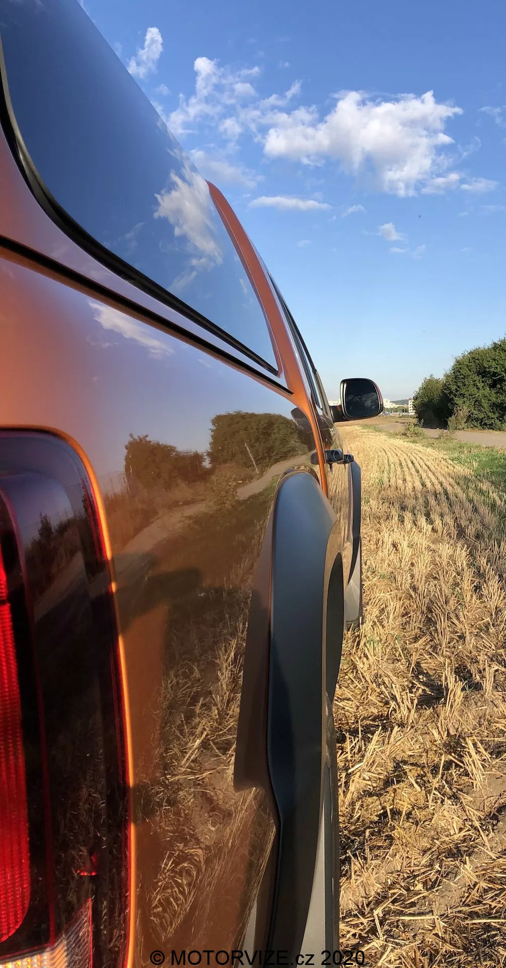 Vista lateral de cerca del panel trasero derecho de un VW Nutzfahrzeuge Amarok 2016, destacando la carrocería metálica brillante y reflectante y la luz trasera integrada con lente clara. La foto, tomada desde un ángulo bajo, muestra la línea de cintura, el borde de plástico negro y el fondo de campo cosechado rural con un cielo azul claro.