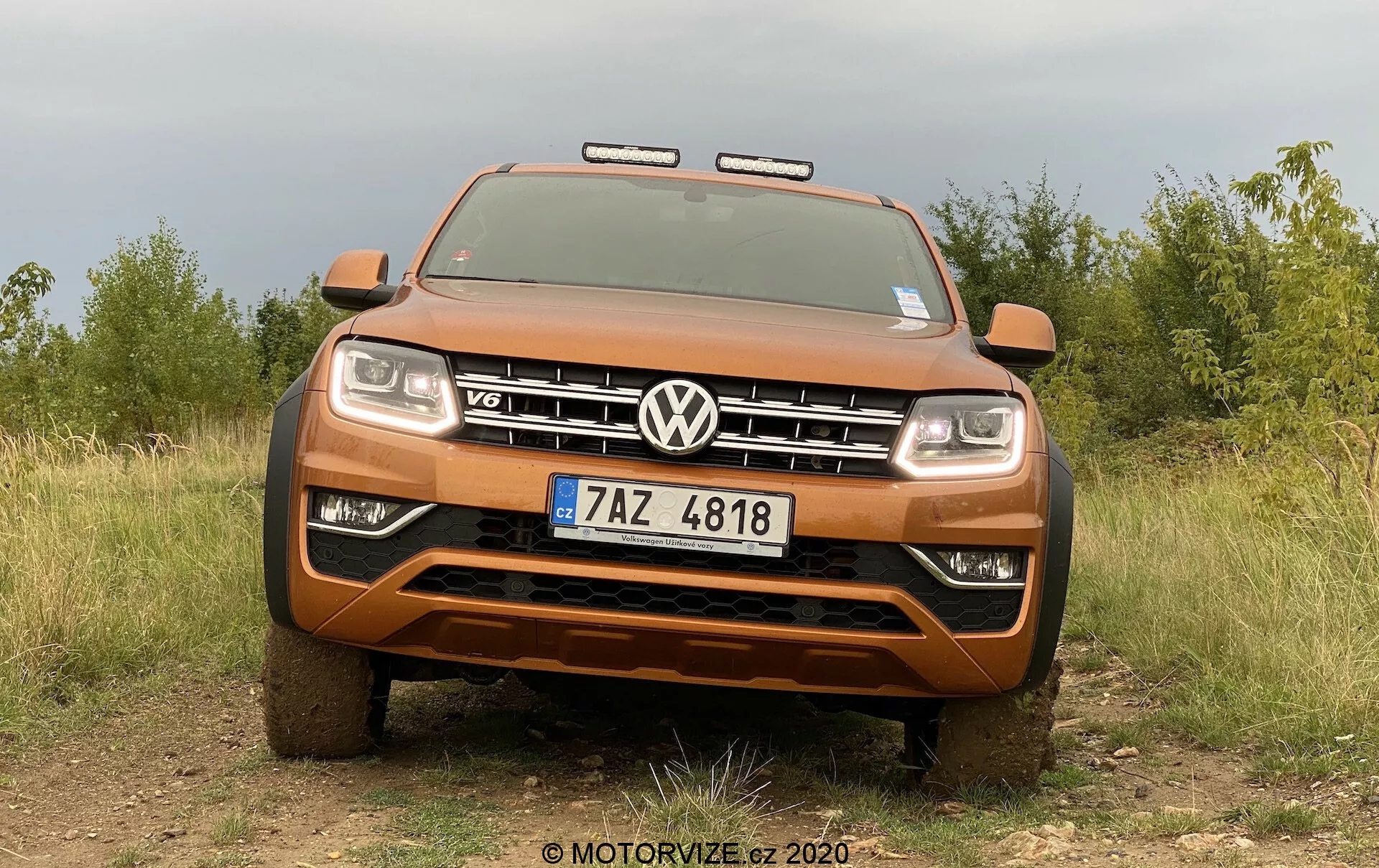 Vista frontal de una camioneta Volkswagen Amarok de color naranja metálico, que muestra su postura robusta y capacidades todoterreno con una parrilla prominente con el emblema VW y la insignia V6. La imagen, tomada desde un ángulo bajo, captura todo el ancho de la parte delantera del vehículo, incluidas las luces delanteras y luces adicionales todoterreno, con un fondo natural de un campo y árboles resaltando la robustez de la camioneta. Las características visibles incluyen un parachoques delantero resistente, placa protectora, sistema de iluminación avanzado activado y matrícula de la República Checa.