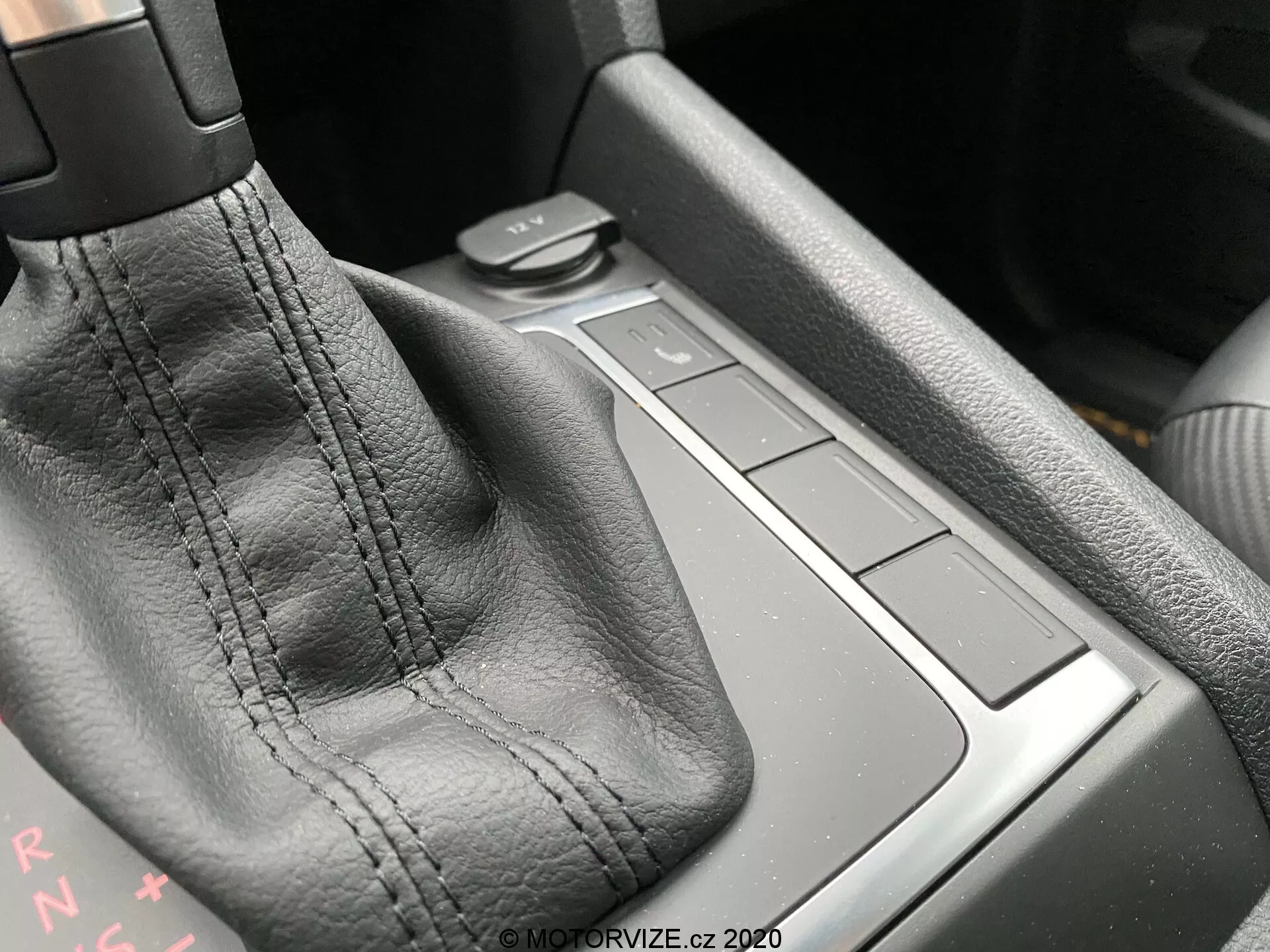 Imagen en primer plano de la consola central del VW Nutzfahrzeuge Amarok 2016, centrada en la zona de la palanca de cambios. La palanca de cambios, cubierta de cuero negro con costuras visibles, es el enfoque principal. Las letras 'PRNDS' detrás de la palanca indican una transmisión automática. A la derecha hay cuatro botones cuadrados a ras de la superficie de la consola, posiblemente controlando varias funciones del coche. El área detrás de los botones puede ser un pequeño espacio de almacenamiento. Los materiales y la calidad de construcción sugieren un alto nivel de artesanía. La luz ambiental desde la esquina superior izquierda resalta los detalles.