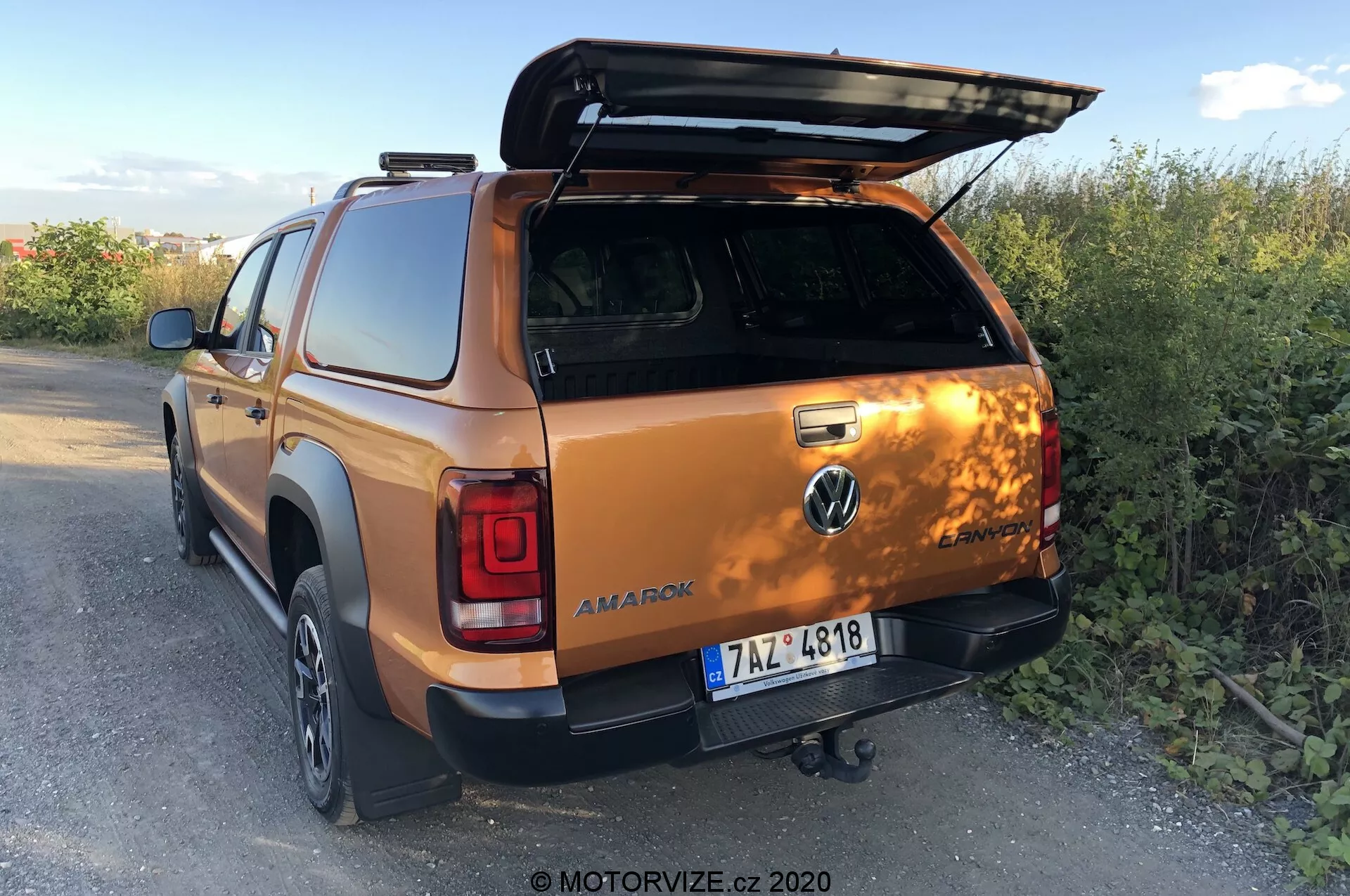 Vista trasera de tres cuartos de un Volkswagen Amarok 2016 en color naranja metálico, destacando la tapa abierta de la cama, el enganche de remolque, las puertas de la cabina doble de la segunda fila y parte de la rueda y el parachoques traseros derechos, situado en un entorno rural al atardecer. Foto de MOTORVIZE.cz, 2020.