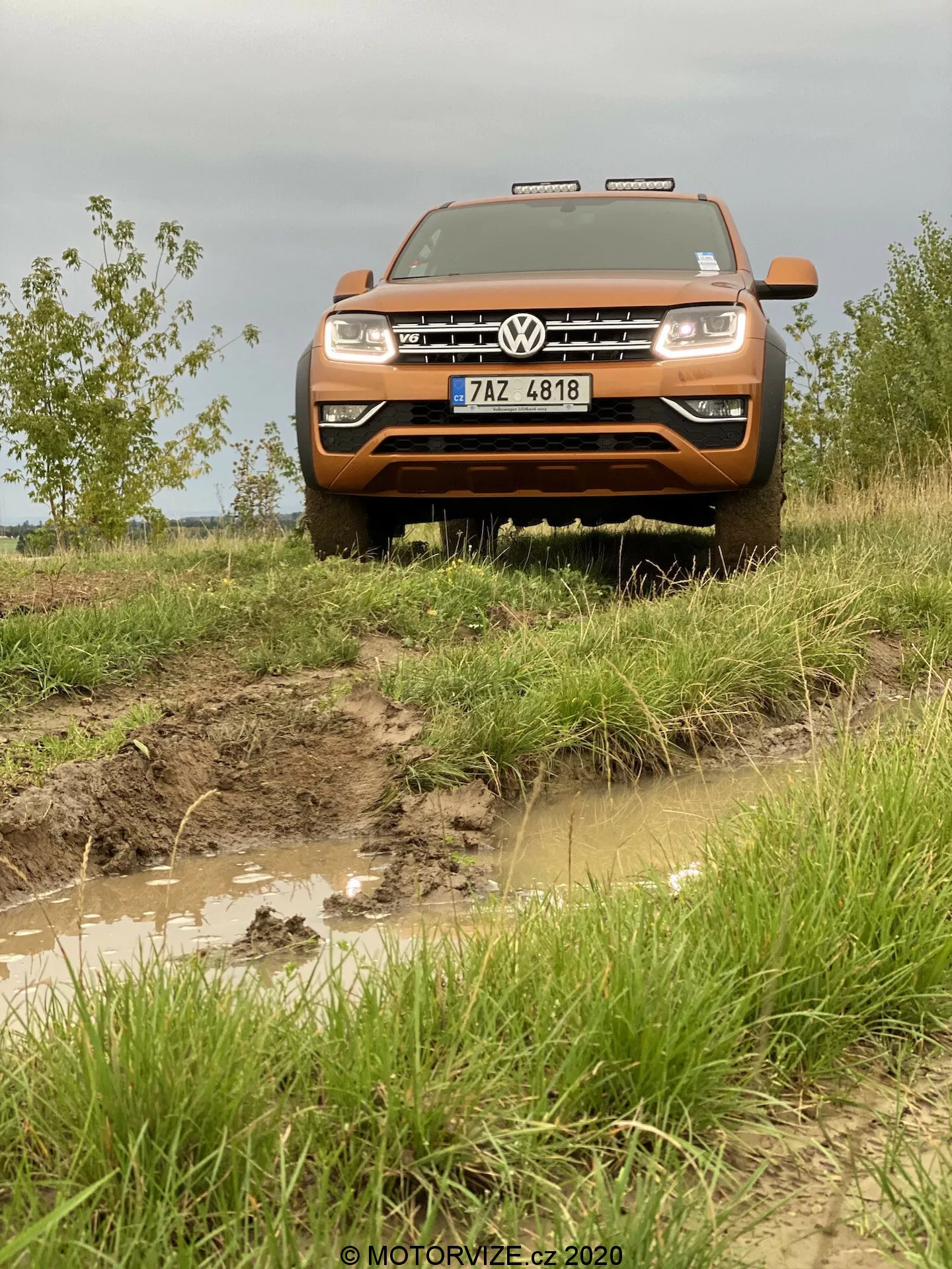 Vista frontal de un vehículo todoterreno Volkswagen Amarok de 2016 en color naranja, destacando la parrilla frontal, el parachoques, el capó y el conjunto de faros. Fotografiada desde un ángulo bajo para resaltar su postura elevada, la imagen revela un sendero accidentado y lodoso con charcos de agua y un exterior salpicado de barro, enfatizando sus capacidades todoterreno. El vehículo cuenta con la parrilla Volkswagen prominente con el emblema, faros rectangulares y luces auxiliares sobre la parrilla. La matrícula es visible en el parachoques delantero robusto, mientras que el capó plano y alargado subraya el tamaño y la potencia del Amarok.