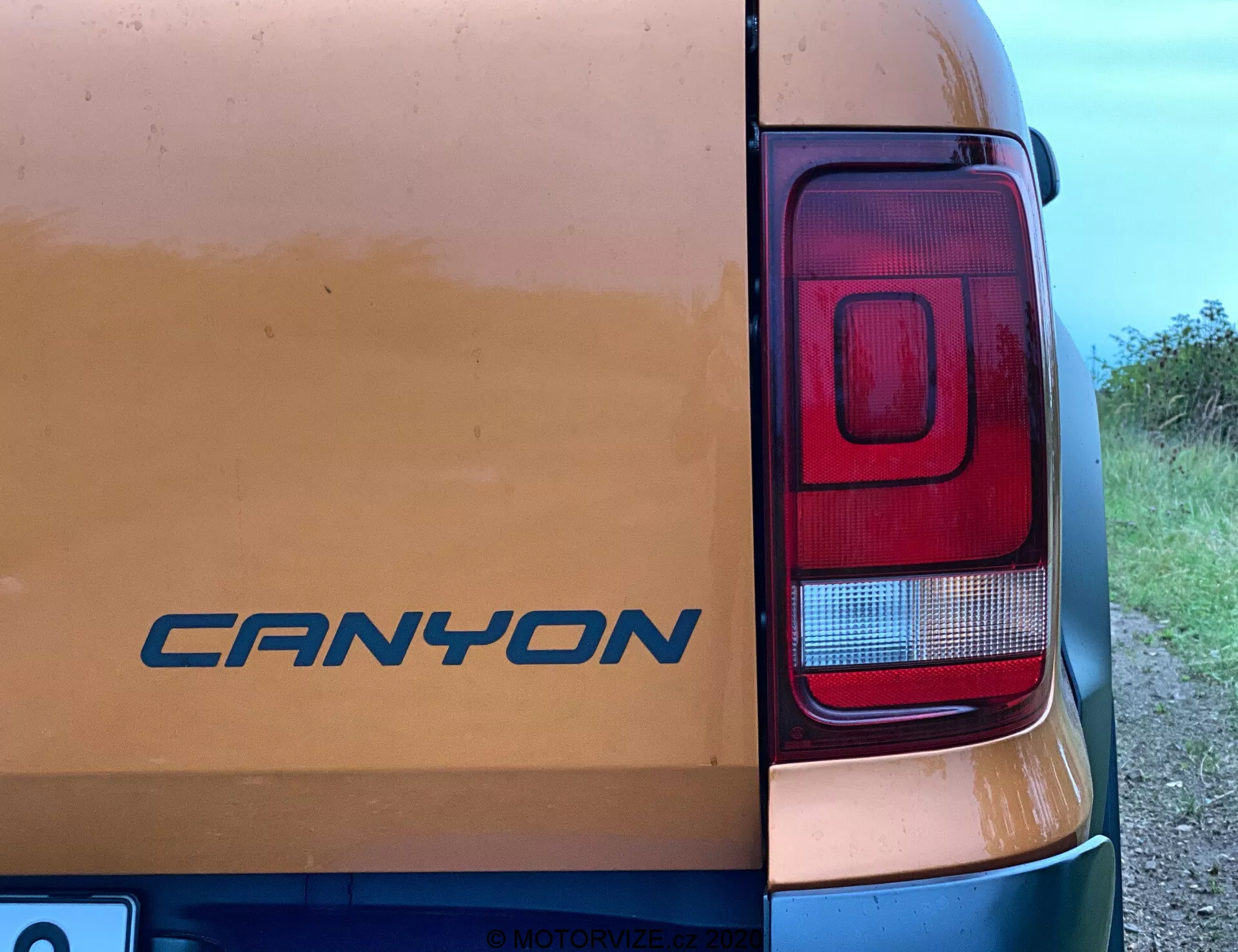 Vista de cerca de la esquina trasera derecha de un vehículo VW Nutzfahrzeuge Amarok 2016. El emblema 'CANYON' es visible en la puerta trasera entre señales de ligera suciedad, sugiriendo uso en caminos sin pavimentar. La fotografía destaca principalmente el conjunto de luces traseras rectangulares y modernas con secciones para freno, reversa y posiblemente señales de giro. Tomada desde un ángulo lateral aproximado de 45 grados, la imagen captura parcialmente el cuerpo del vehículo con un fondo rural o semi-rural.