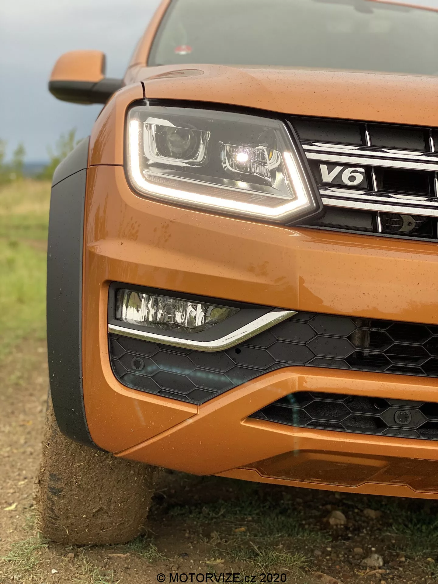 Primer plano de la esquina delantera izquierda de un VW Nutzfahrzeuge Amarok 2016 naranja mostrando faro con lente claro moderno y tecnología LED, luces antiniebla en bisel negro, parrilla parcialmente visible con insignia V6, parachoques delantero con malla de panal negra, diseño contorneado del capó y rueda grande parcialmente visible, con un fondo borroso que enfatiza la textura y el diseño robusto del coche