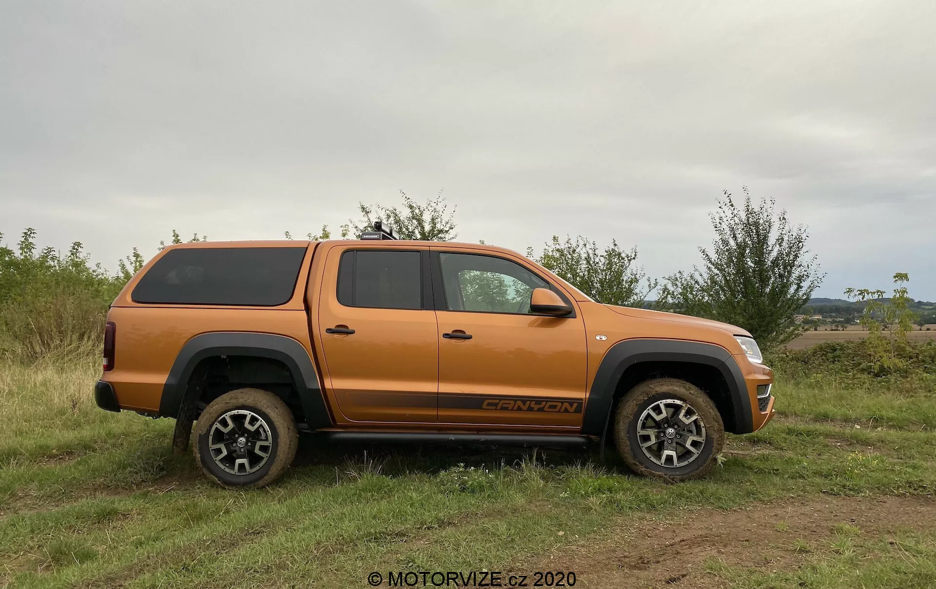 Perfil lateral de una camioneta pickup VW Nutzfahrzeuge Amarok 2016 naranja con una cubierta de tonel rígida, estacionada en terreno irregular y con hierba. El vehículo presenta una alta distancia al suelo, ruedas y neumáticos robustos, y un diseño de aleación con múltiples radios adecuado para su uso todoterreno. Los elementos visibles incluyen espejos laterales, manijas de las puertas, arcos de rueda, estribo, cabina espaciosa de cuatro puertas, insignia en la rueda delantera y texto de nivel de acabado sobre la rueda trasera. La luz natural y el ángulo nivelado destacan los detalles del camión sin sombras fuertes, enfatizando su capacidad todoterreno y almacenamiento seguro de carga.