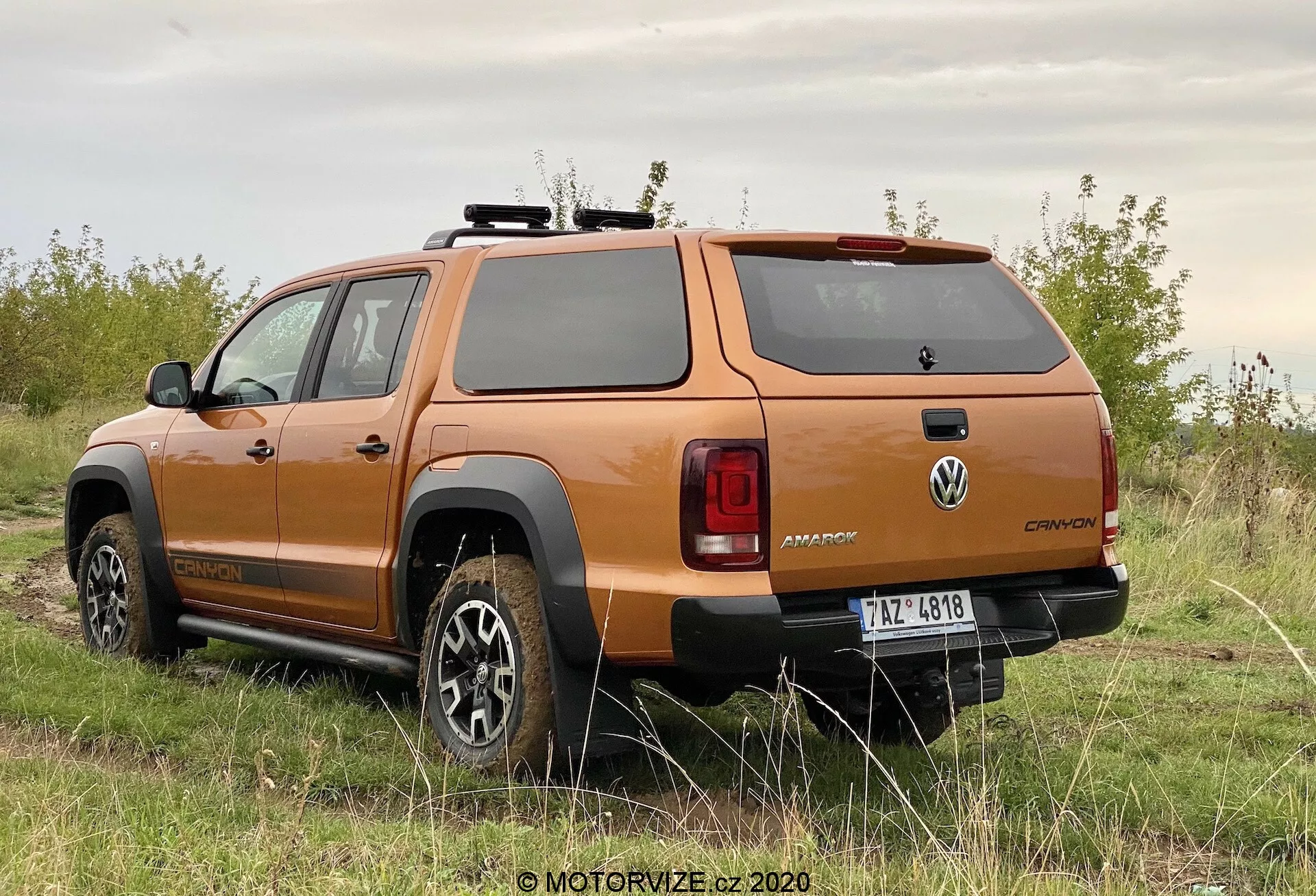Vista trasera de tres cuartos de un camión pickup Volkswagen Amarok 2016 naranja, mostrando el diseño de las ruedas, el perfil lateral, la ventana trasera, las luces traseras, el parachoques trasero robusto y la cubierta rígida de la cama, estacionado en una superficie sin pavimentar con vegetación, destacando sus capacidades todoterreno.