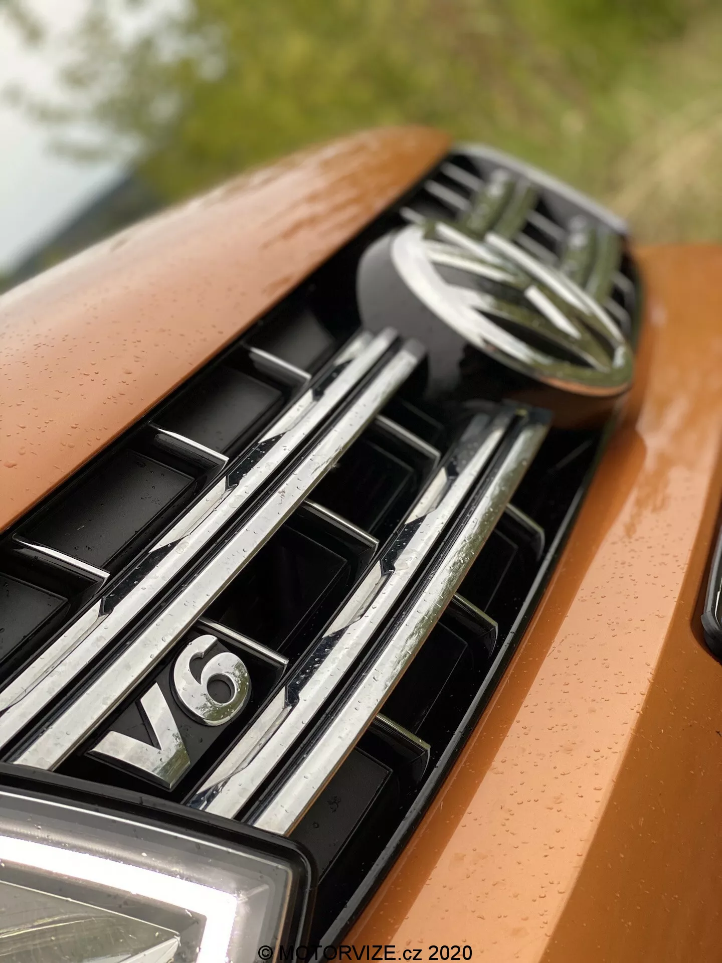 Plano detalle del capó y la parrilla de un coche, centrado en la parte superior que muestra un emblema central y una insignia del motor V6. La parrilla cuenta con lamas horizontales reflectantes de cromo, indicativas de un vehículo de lujo. La pintura del coche es de un color naranja o bronce metálico con una iluminación suave y difusa que acentúa su diseño suave y sensación premium.