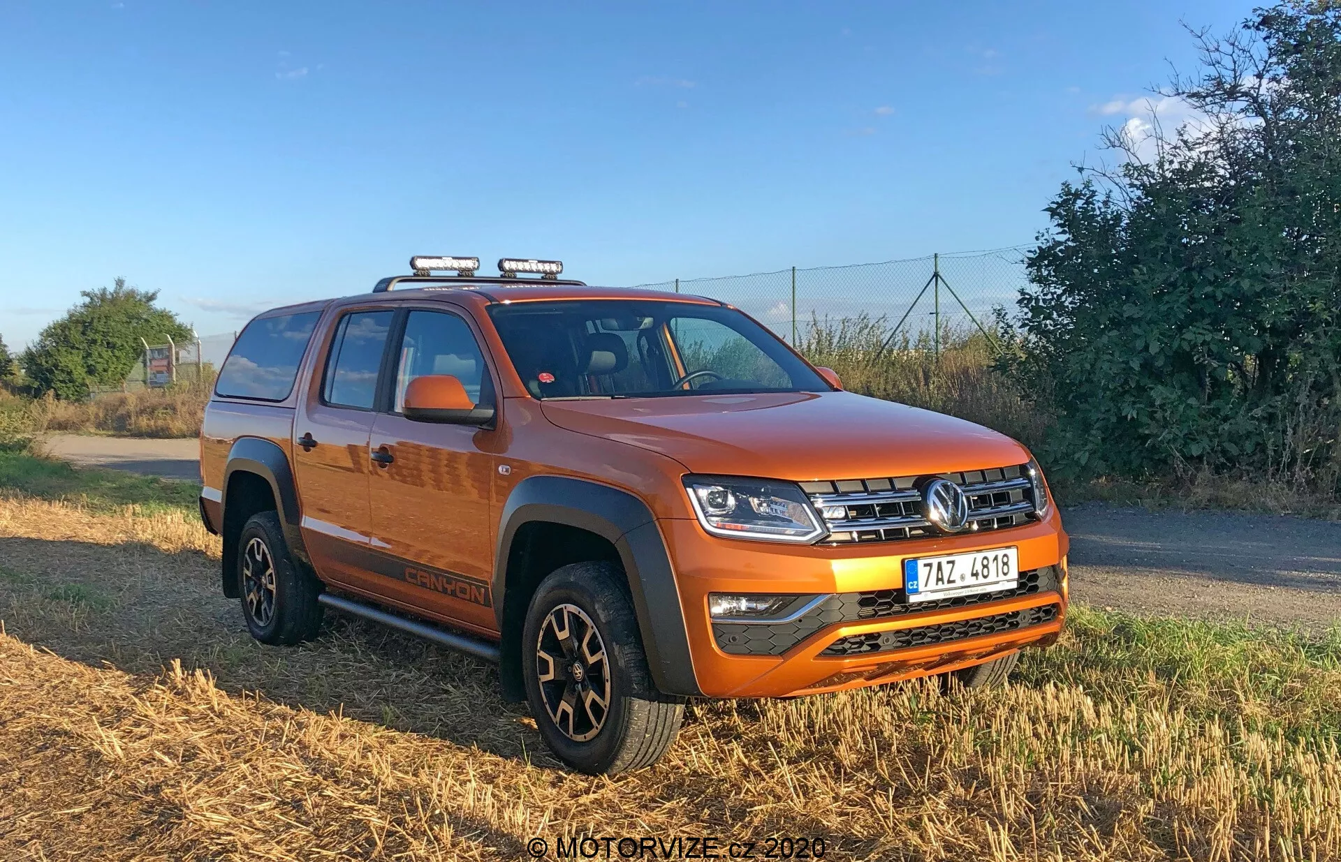 Imagen de una vibrante camioneta VW Nutzfahrzeuge Amarok 2016 de cuatro puertas en color naranja tomada en ángulo de tres cuartos, mostrando el frente y el costado del vehículo. La imagen resalta su parrilla frontal con diseño horizontal, faros LED complejos, luces auxiliares montadas en el techo, parachoques sólido con faros antiniebla, placa protectora, sección central elevada del capó, protectores negros en los arcos de las ruedas, llantas negras con neumáticos multipropósito, estribos, espejos con indicadores integrados, cama de camión cubierta con una lona y rieles en el techo para almacenamiento adicional.