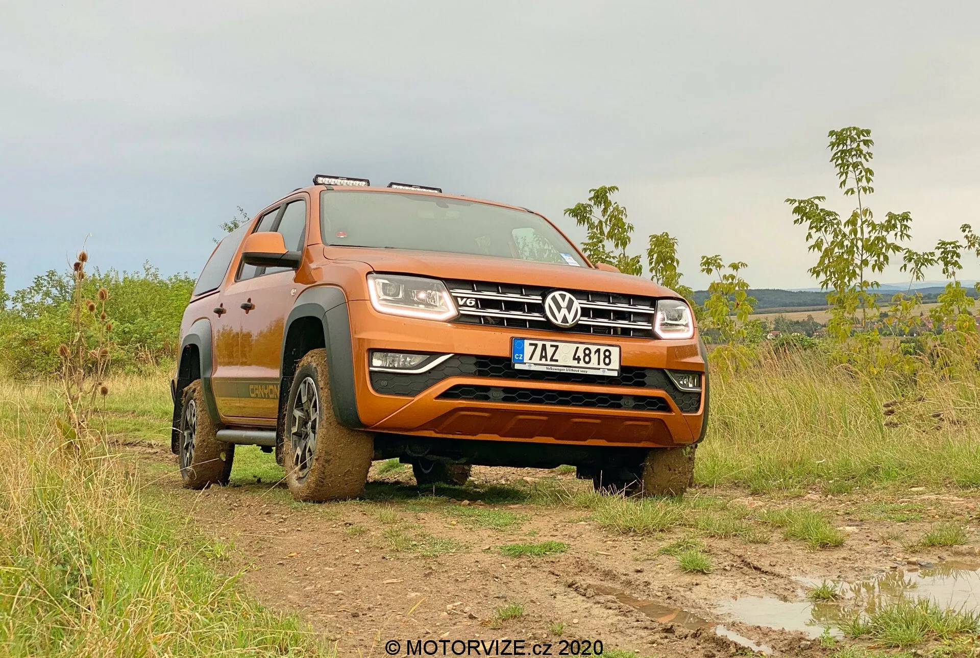 Ángulo delantero de tres cuartos bajo de un Volkswagen Amarok 2016 en un campo embarrado, mostrando su pintura naranja, parrilla delantera con el emblema de Volkswagen, faros, diseño de ruedas y capacidad todoterreno. La robustez del vehículo se resalta con el barro en los neumáticos y los lados inferiores, con las ruedas delanteras giradas hacia la izquierda, destacando los patrones de la banda de rodadura y la altura de conducción. Ambientado en un fondo suavemente iluminado, ya sea por la mañana o a última hora de la tarde, la imagen subraya la utilidad todoterreno del Amarok y su avanzado sistema de iluminación. La matrícula es visible y debe ser ocultada para la privacidad.