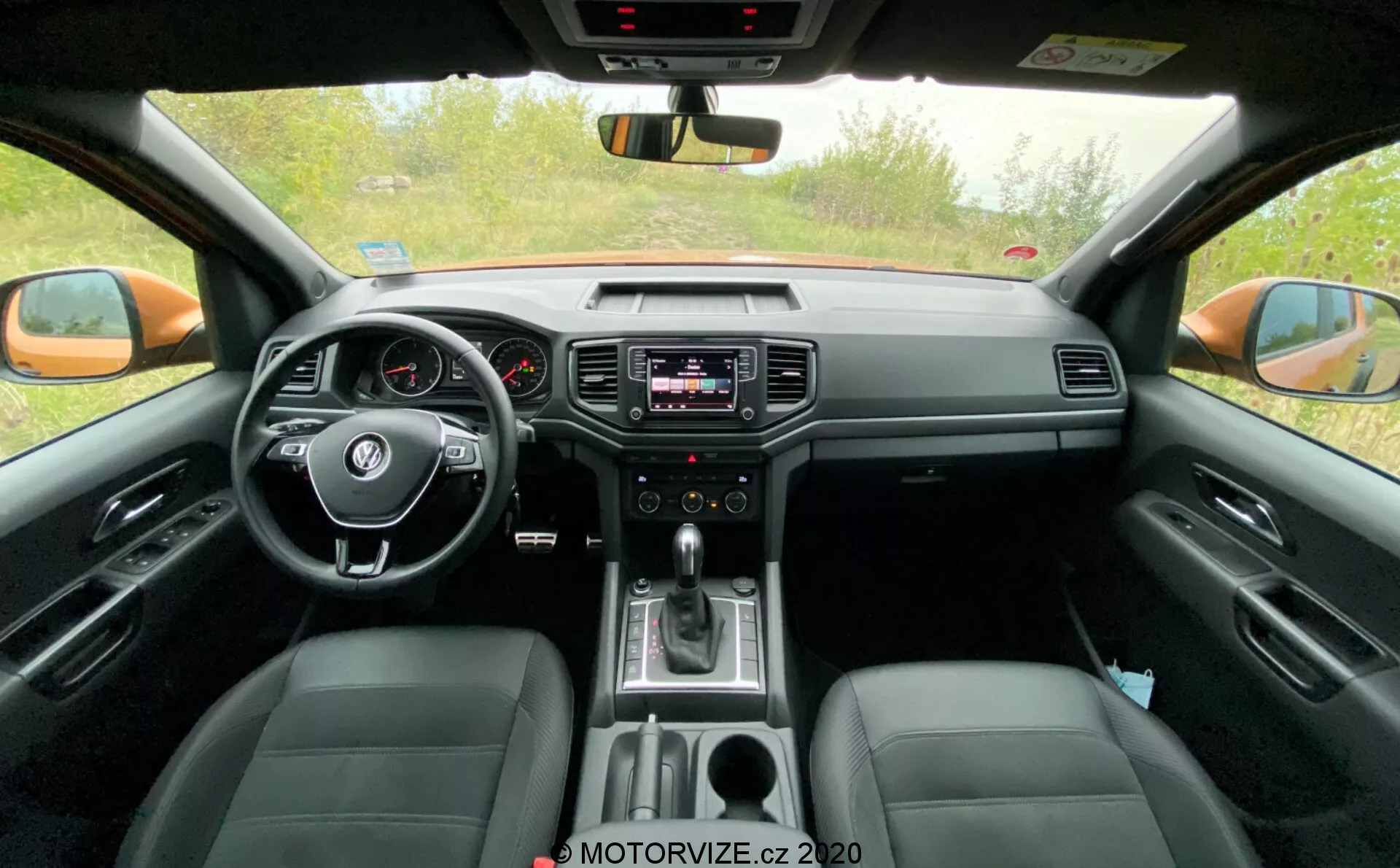 Vista delantera del habitáculo del VW-Nutzfahrzeuge Amarok de 2016. Tomada desde la perspectiva del asiento trasero, esta imagen muestra el volante central con controles integrados, el cuadro de instrumentos analógico con una pequeña pantalla digital, el sistema de infoentretenimiento en el centro del tablero con controles de audio y clima, la palanca de cambios indicando transmisión automática y asientos de cuero. La imagen también captura el espejo retrovisor, los espejos retrovisores exteriores y las ventanas laterales frontales, destacando tanto las características tecnológicas como de confort.