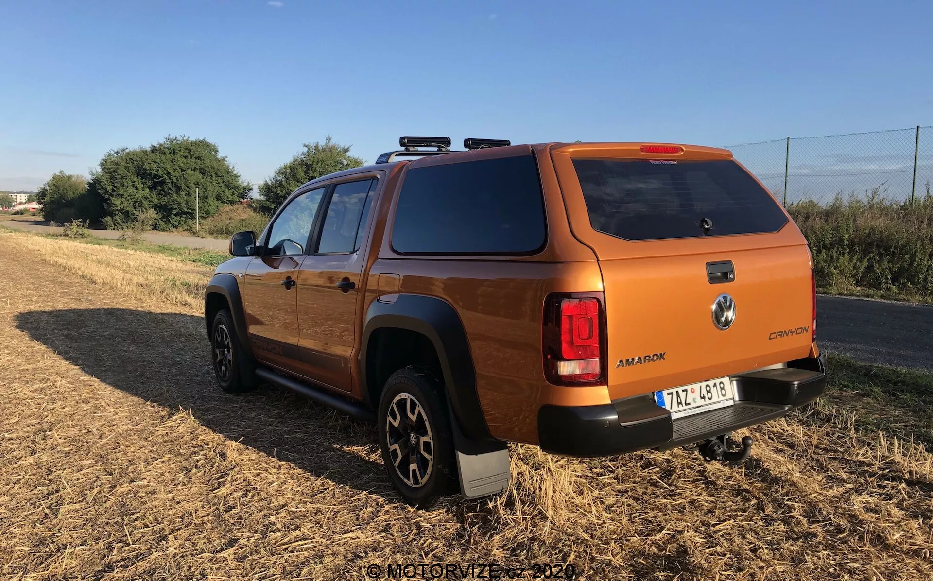 Vista trasera de un vehículo utilitario o todoterreno moderno de color bronce o naranja quemado, posiblemente un SUV mediano o una camioneta pick-up, con una cubierta en la caja de carga. El ángulo resalta la parte trasera y el perfil lateral, incluyendo la rueda trasera y la puerta del pasajero, situado en un terreno sin pavimentar con vegetación al fondo. El vehículo exhibe un diseño robusto y cuadrado típico de vehículos todoterreno, con luces traseras envolventes, neumáticos grandes para todo terreno en llantas de aleación negras, emblemas visibles, rieles en el techo, cristales traseros tintados y una cubierta sólida en la caja de carga.