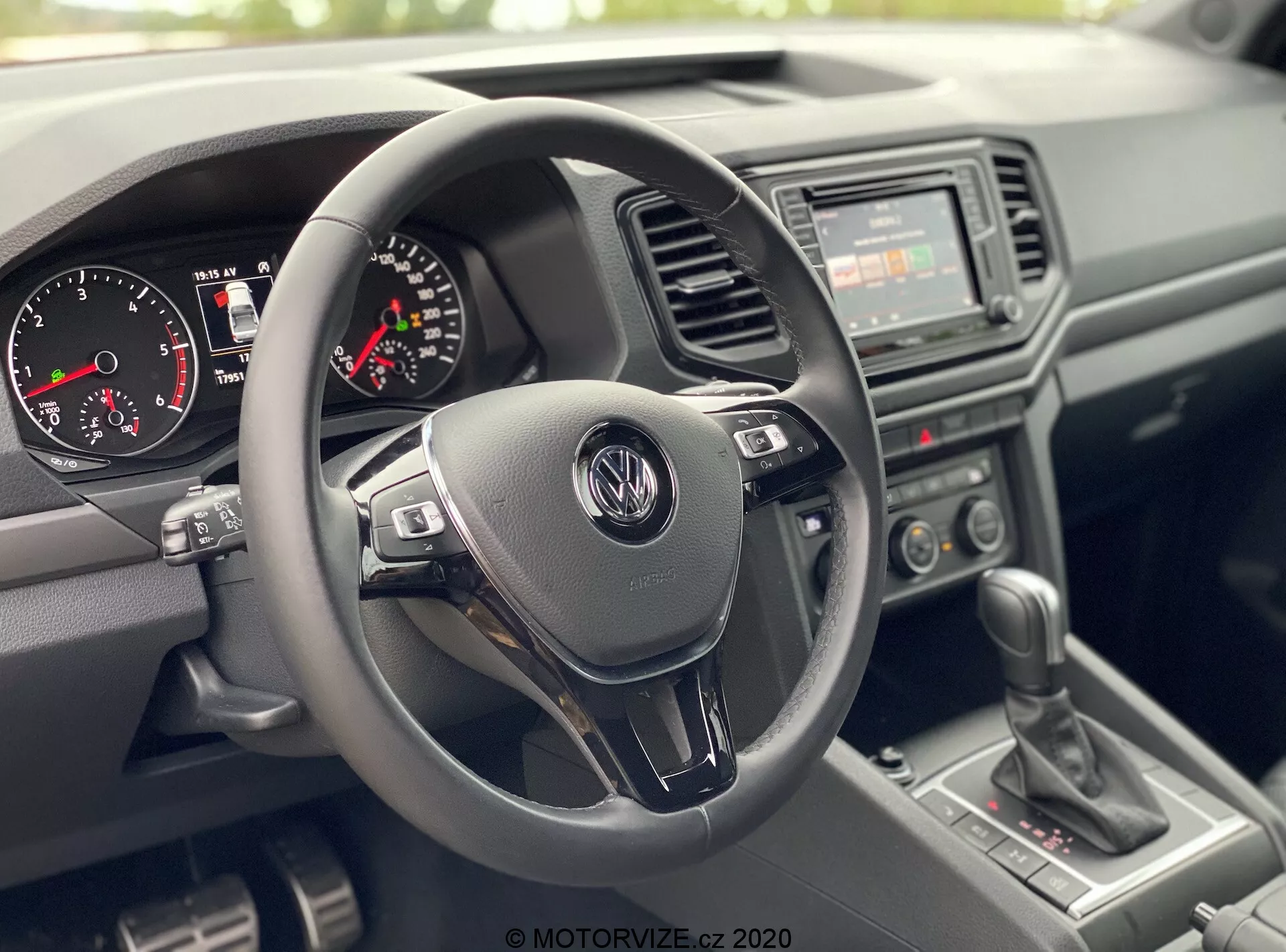 Vista del interior del VW Nutzfahrzeuge Amarok 2016 desde la perspectiva del pasajero delantero, mostrando el área del puesto de conducción. Enfoque central en un volante de tres radios con logotipo de Volkswagen y botones de control multifuncional, clúster de instrumentos con velocímetro analógico, tacómetro y pequeña pantalla digital, radio básico con interruptores de control climático manual en la consola central, palanca de cambios manual y materiales plásticos texturizados para el salpicadero y las puertas, destacando un diseño duradero y práctico.