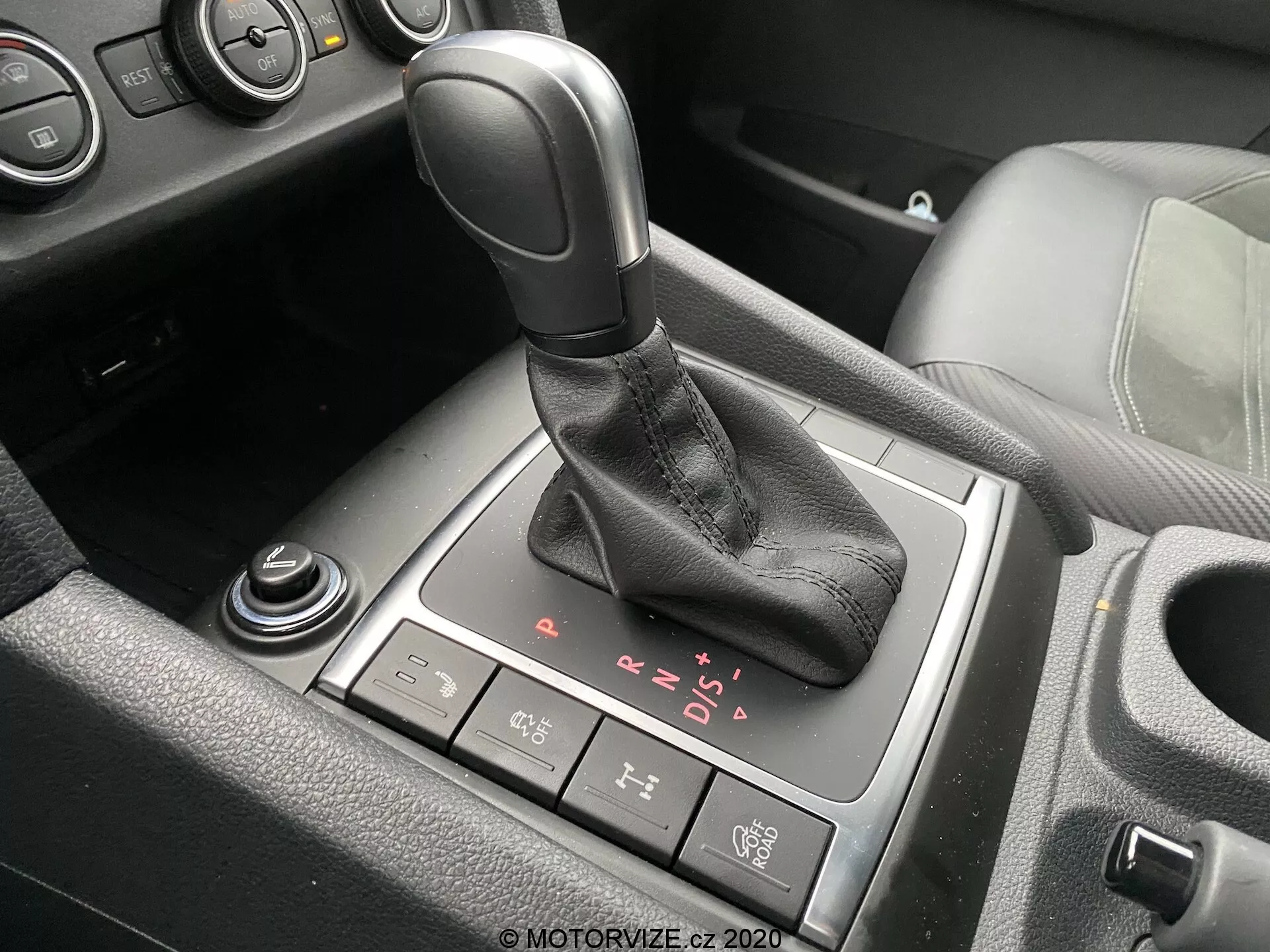 Vista detallada de la zona de la palanca de cambios en el interior del VW Nutzfahrzeuge Amarok 2016. La palanca de cambios automática, con un mango de color negro y una funda de cuero, está posicionada en el centro de la imagen con opciones visibles de P, R, N, D y modo secuencial manual. Detrás de la palanca de cambios se encuentra un botón de freno de estacionamiento electrónico, mientras que un dial giratorio parcialmente visible y otro dial están situados al frente y en la parte inferior izquierda, respectivamente. Vistas parciales del portavasos y la consola central con acentos de plástico negro y aluminio o cromo también están presentes, indicando un vehículo moderno con un acabado interior de nivel medio o superior.