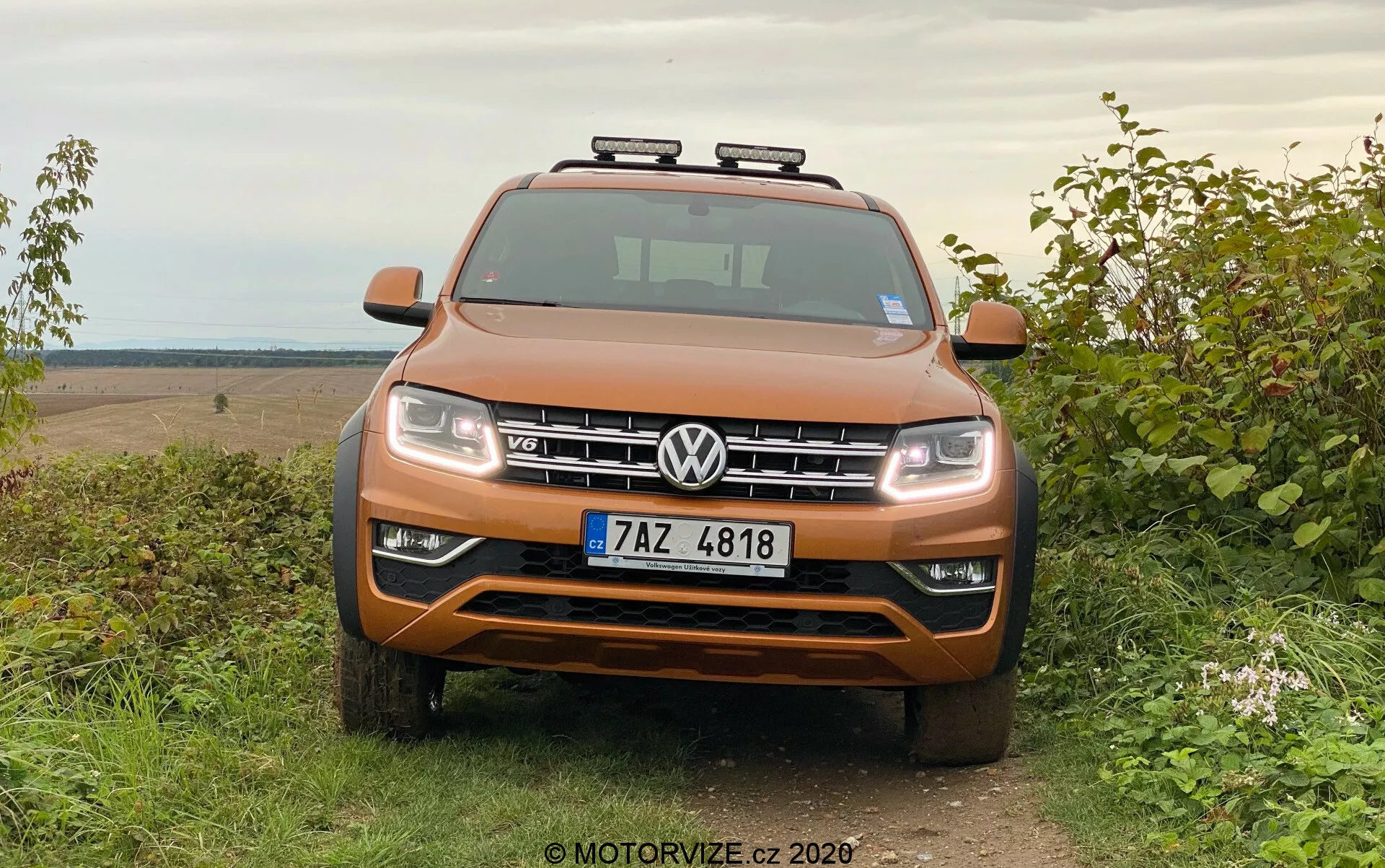 Vista frontal de un SUV Volkswagen Amarok de color naranja metálico estacionado en el césped con un fondo de configuración rural todoterreno, mostrando una rejilla simétrica, faros, acentos cromados y luces auxiliares en el techo.