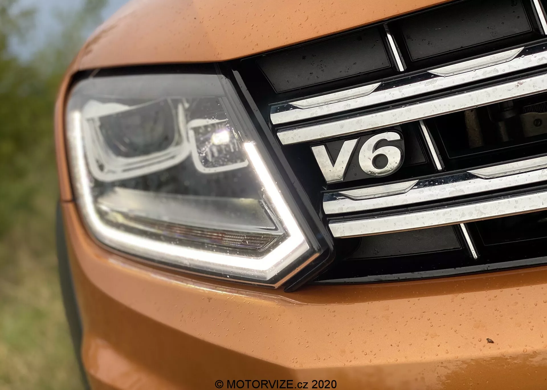 Primer plano de la sección frontal izquierda del VW Nutzfahrzeuge Amarok 2016. La imagen captura un faro moderno y angular con luces diurnas LED y tecnología de haz de proyector, junto con detalles cromados. La parrilla presenta tiras horizontales de cromo y una insignia 'V6', indicando el tipo de motor de seis cilindros del coche. Parte del capó aparece en un tono naranja vibrante, sugiriendo un diseño deportivo.