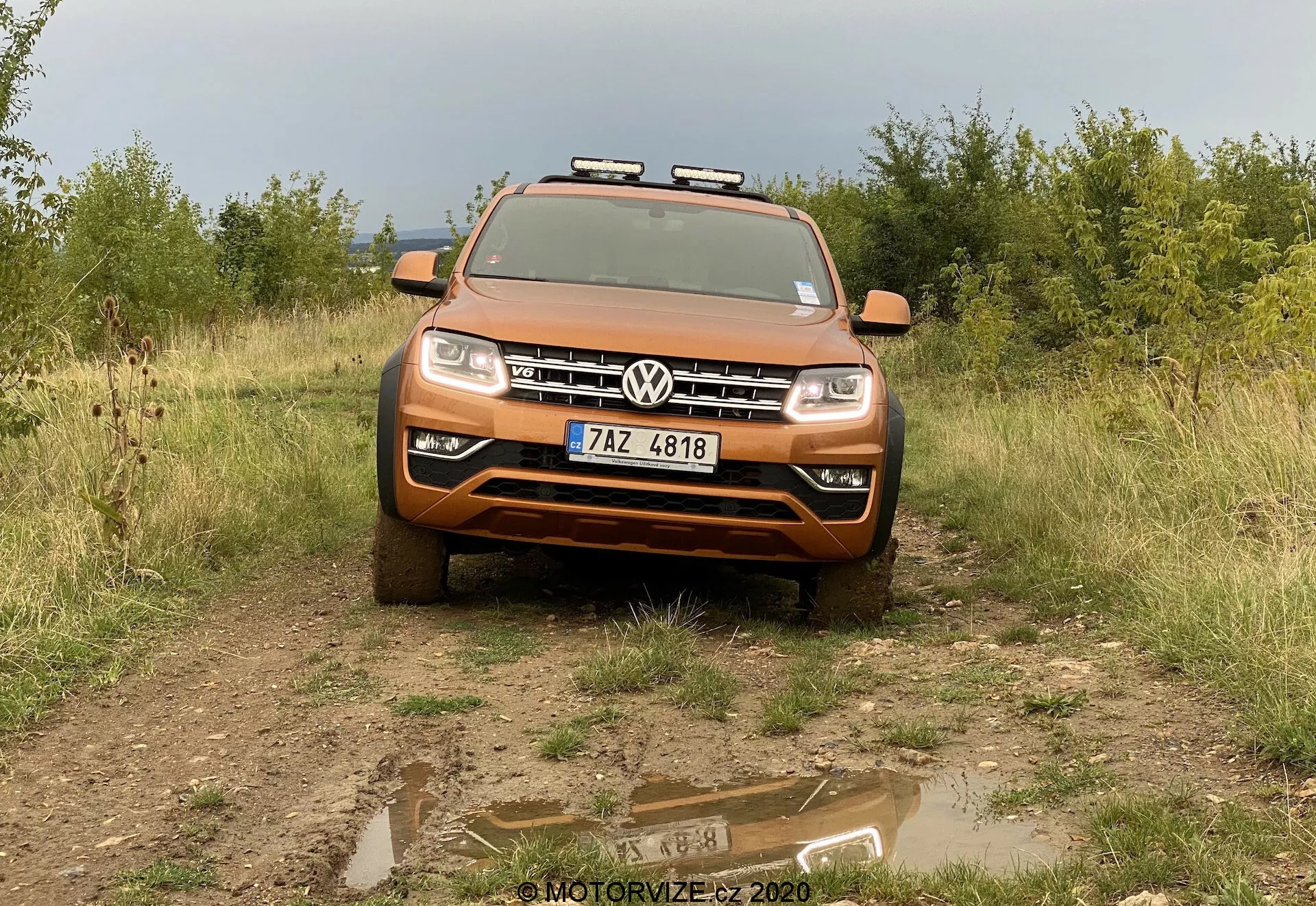 Vista frontal de un Volkswagen Amarok 2016 SUV de color naranja metálico en un terreno no pavimentado con hierba y arbustos en el fondo. El vehículo está orientado hacia la cámara en un ligero ángulo hacia abajo, con un charco de agua poco profundo en primer plano reflejando la imagen del coche. La parrilla delantera presenta el logotipo de Volkswagen en el centro y el capó muestra sus contornos y líneas de diseño. Equipado con faros LED elegantes y luces antiniebla abajo, una placa deslizante de plata en la parte inferior sugiere capacidades todoterreno. Luces auxiliares están montadas en un portaequipajes de techo, mejorando su estética robusta. La placa de matrícula está ubicada en el centro del parachoques delantero. La foto captura el ancho completo del vehículo, mostrando detalles relevantes a la tecnología, diseño y adaptaciones todoterreno.