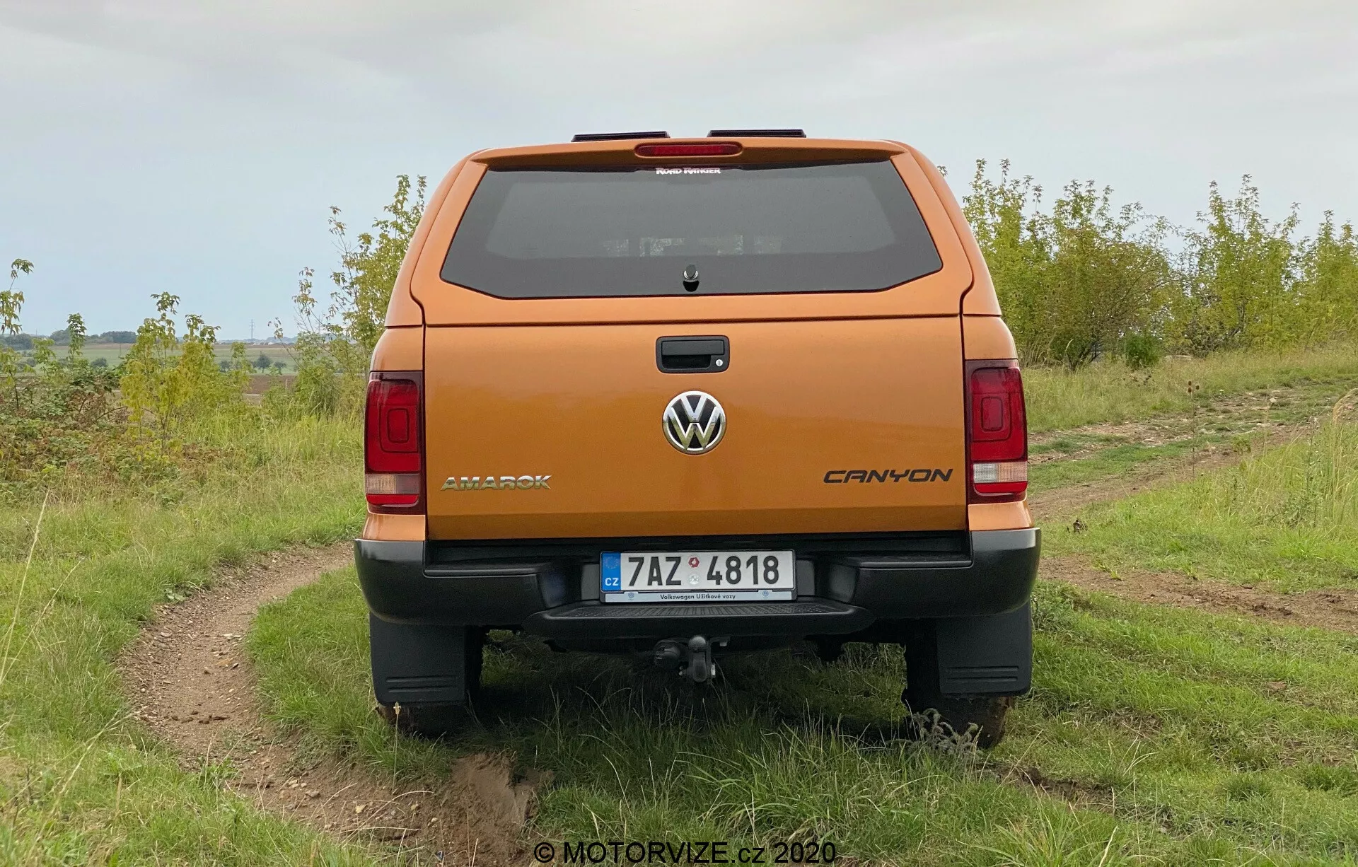 Vista trasera de una camioneta Volkswagen Amarok 2016 de color naranja metálico estacionada afuera en un camino de tierra irregular con hierba a ambos lados, bajo un cielo nublado. La foto muestra una clara perspectiva del portón trasero y las características del diseño trasero, incluyendo el emblema de Volkswagen, la placa de nombre Amarok y el emblema V6. El vehículo tiene un parachoques trasero con un escalón integrado, una placa de matrícula ubicada en el centro y un enganche de remolque visible. Se pueden ver las luces traseras orientadas verticalmente y la tercera luz de freno en la línea del techo por encima del parabrisas trasero, así como las líneas del descongelador.