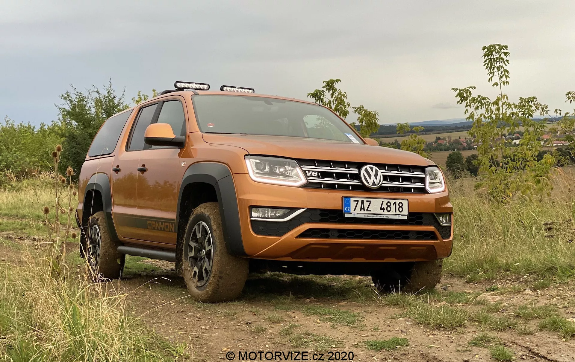 La imagen muestra un SUV moderno, un modelo Volkswagen Amarok 2016, estacionado al aire libre en un terreno irregular que demuestra su capacidad todoterreno. Visto desde un ángulo frontal bajo, se aprecia un cuarto delantero del vehículo cubierto de barro debido a su reciente uso todoterreno. La amplia parrilla delantera con el distintivo emblema VW está flanqueada por faros rectangulares con luces diurnas LED. Bajo la parrilla, un robusto parachoques delantero con una placa protectora de plata está diseñado para proteger los bajos del vehículo. El capó contorneado añade una apariencia musculosa al SUV. Las barras de techo son visibles, indicando capacidad adicional de carga. Las ruedas, parcialmente cubiertas de barro, tienen neumáticos todoterreno de gran tracción. Un prominente arco sobre la rueda delantera y un estribo lateral facilitan la entrada y salida del vehículo. También se distinguen las luces del techo y los espejos laterales. El coche se encuentra en un fondo de vegetación y terreno montañoso bajo un cielo nublado, enfatizando su atractivo todoterreno y al aire libre. El volante a la izquierda y la matrícula visible sugieren un país de conducción por la derecha. Esta toma destaca el diseño del coche, las características todoterreno y las avanzadas tecnologías de iluminación.