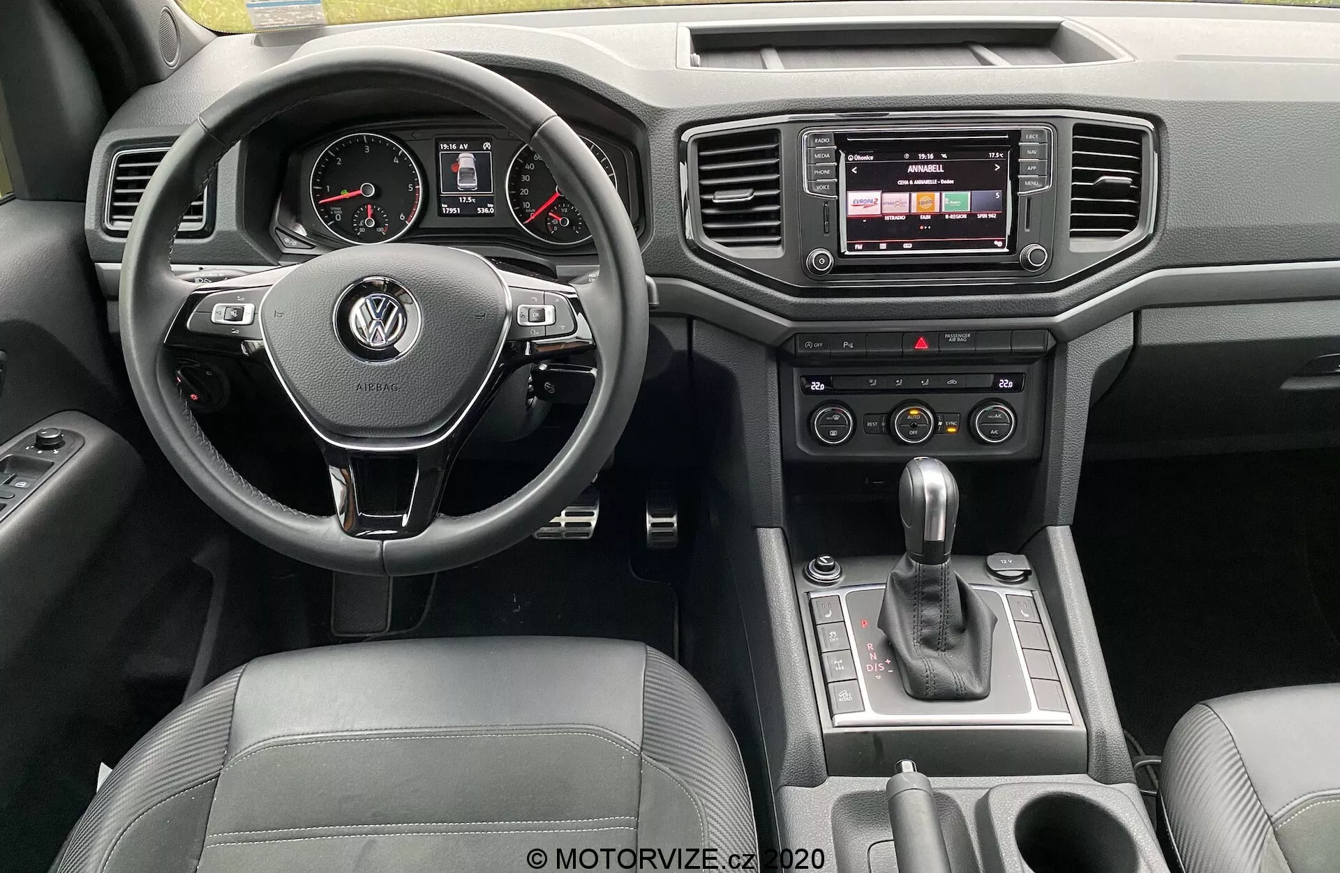 Vista del tablero de instrumentos del Volkswagen Amarok 2016 desde la perspectiva del conductor. El volante multifuncional envuelto en cuero con el logo de VW está en el centro, con diales analógicos y una pantalla digital en el panel de instrumentos. La consola central cuenta con un sistema de infoentretenimiento de pantalla táctil, botones de control climático y una palanca de cambios automática. Detalles de lujo incluyen acabados en cuero y cromo, con controles de la puerta a la vista.