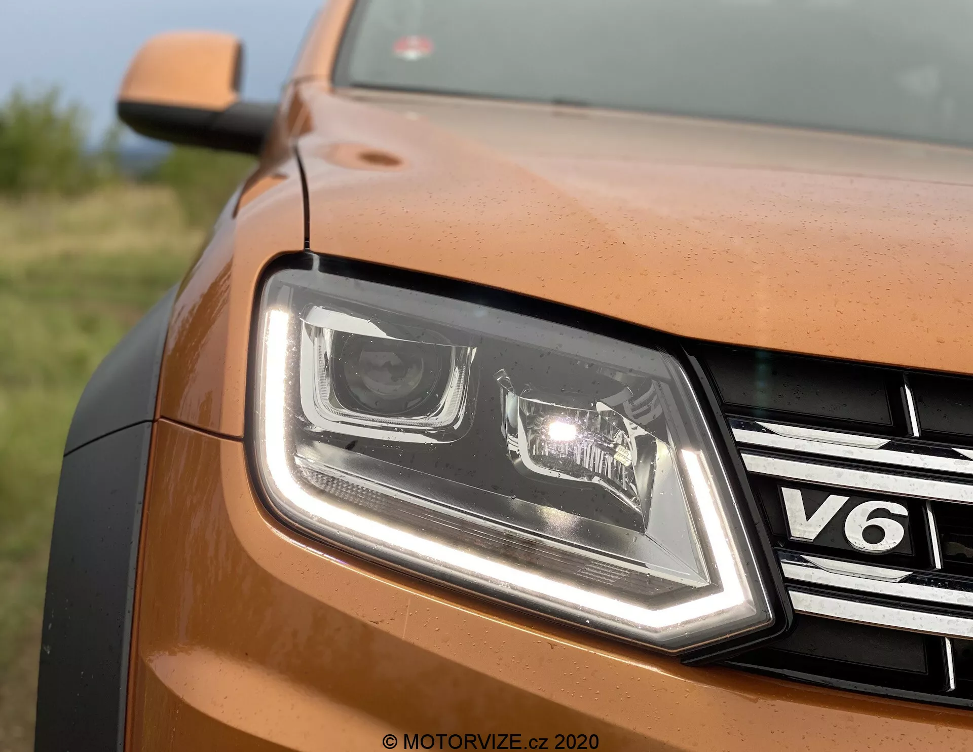 Vista frontal parcial en primer plano del lado delantero izquierdo de un VW Nutzfahrzeuge Amarok 2016 de color bronce o cobre. La imagen muestra el diseño de los faros LED con luces diurnas distintivas, una parrilla cromada con el emblema 'V6', panel de carrocería de pintura metálica, borde contorneado del capó y la marca de agua 'MOTORVIZE.CZ 2020', lo que sugiere origen del sitio de reseñas checo.