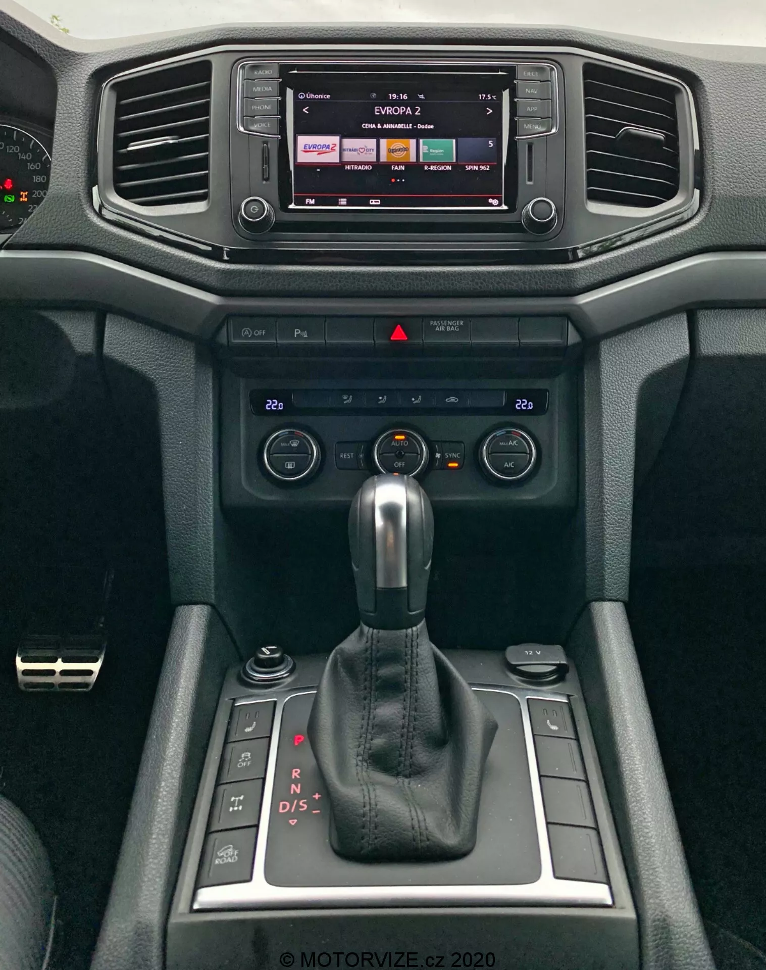 Vista detallada de la consola central moderna del VW Amarok 2016, destacando el sistema de infoentretenimiento y la palanca de cambios. La pantalla de infoentretenimiento indica capacidades táctiles, probablemente mostrando salida de radio o medios. Debajo están las salidas de aire horizontales rodeadas de materiales suaves al tacto con un diseño texturizado. Los controles de clima y los interruptores se encuentran debajo de las rejillas de ventilación para ajustar la temperatura y el flujo de aire. Una palanca de cambios automática en modo 'P' domina el centro, indicando la transmisión automática del vehículo con opciones de cambio manual. Compartimentos pequeños están junto a la palanca de cambios, junto con un dial giratorio, probablemente para el control de infoentretenimiento o modos de conducción. El diseño interior enfatiza tanto la funcionalidad como la comodidad.
