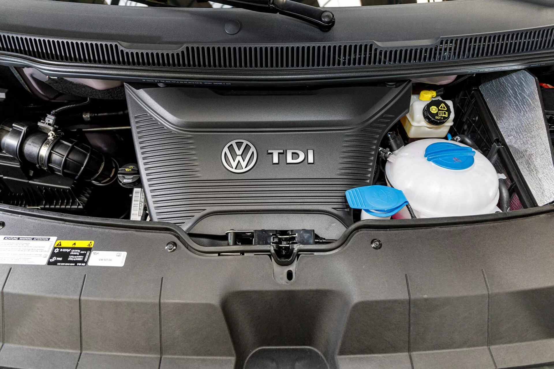 Vista cercana del compartimento del motor diésel de VW Nutzfahrzeuge Transporter 2019 (T6.1 California) mostrando la cubierta del motor TDI con el logotipo de Volkswagen, el depósito de refrigerante y la batería del automóvil