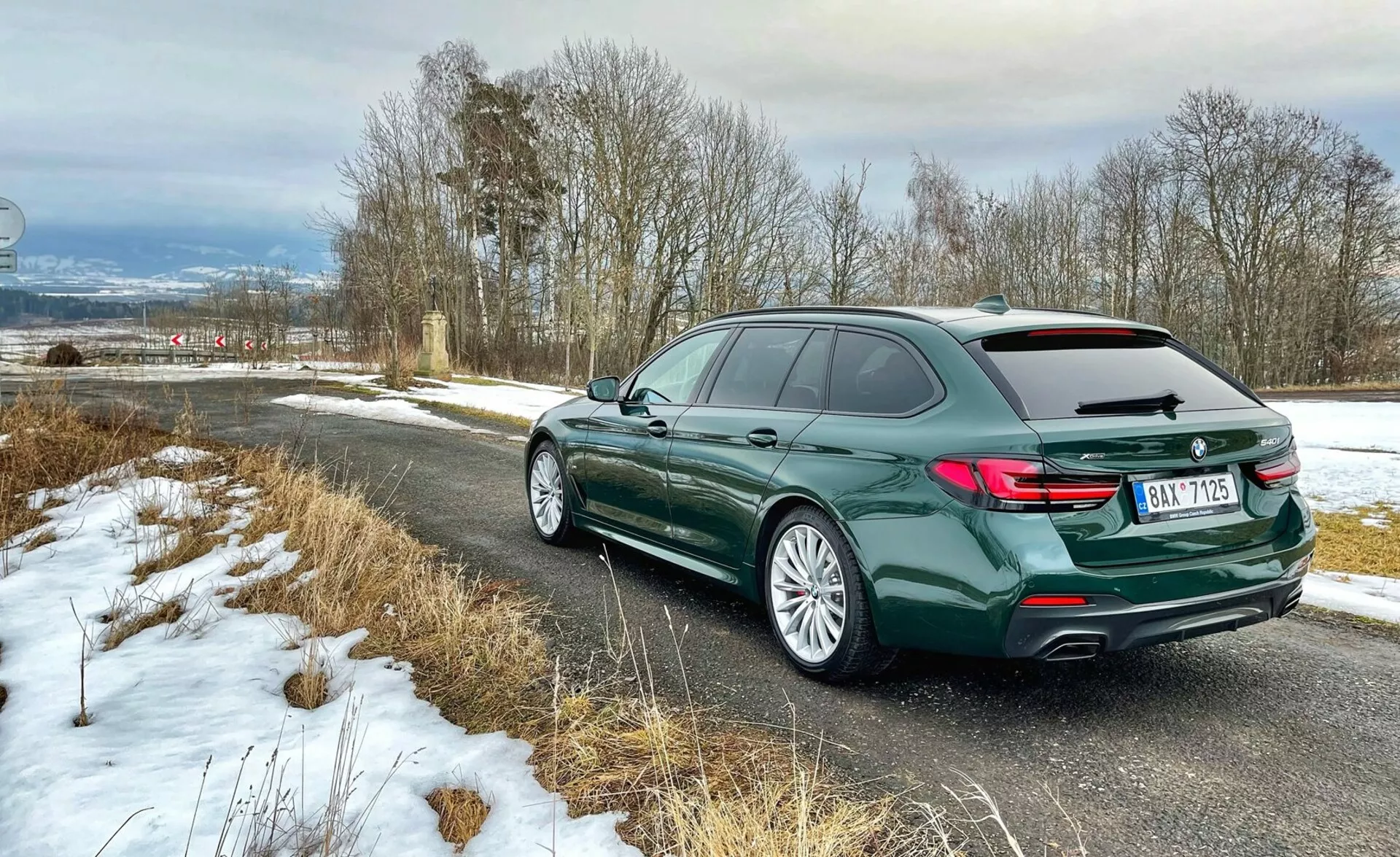 Vista trasera en tres cuartos de un familiar BMW, mostrando la parte trasera del vehículo con luces traseras en forma de L, el perfil lateral izquierdo incluyendo puertas y la curvatura del techo con barras en el techo, llantas de aleación con neumáticos de carretera, en pintura verde oscuro metálico, estacionado en un camino rural con nieve ligera, destacando elementos de diseño de lujo y moderno.