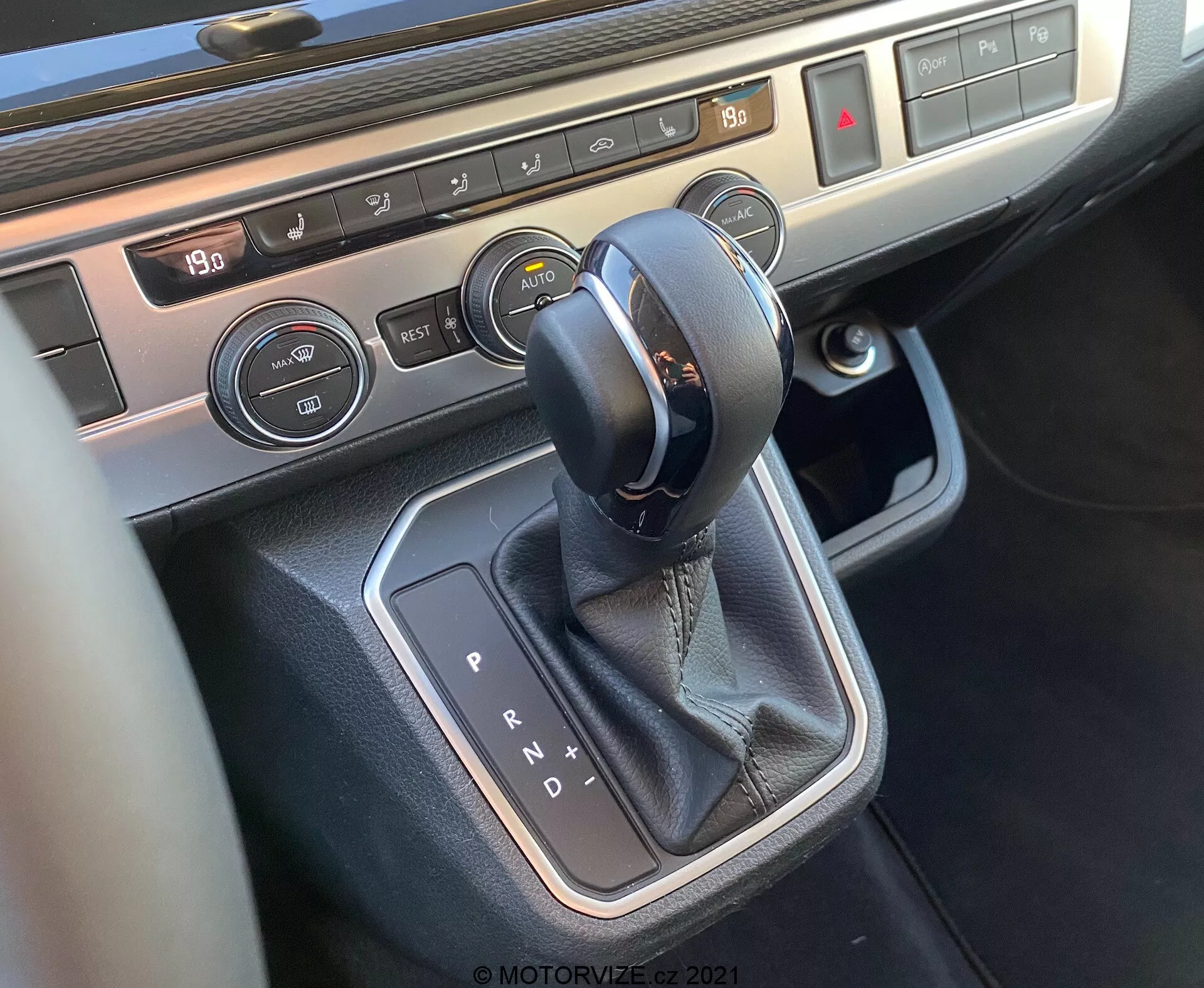 Vista de primer plano de la consola central del VW Nutzfahrzeuge T6-1 Multivan 2019 centrada en el área de la palanca de cambios. La imagen muestra un selector de transmisión automática con costuras de cuero, rodeado por un adorno metálico, botones de control del clima con pantalla de temperatura y controles de calefacción de asientos, destacando la tecnología interior moderna y los materiales premium del vehículo.
