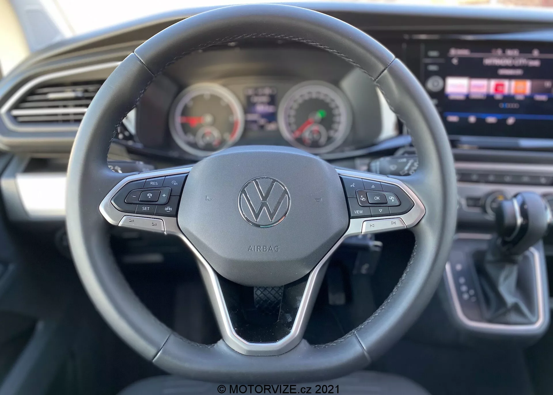 Vista interior de un VW Nutzfahrzeuge Transporter 2019 (T6-1 Multivan) centrada en la perspectiva del conductor. Vista detallada del volante con el logotipo de Volkswagen y botones de control para varias funciones como control de crucero y navegación de sistemas multimedia. Detrás del volante, el panel de instrumentos muestra un velocímetro y un tacómetro de estilo analógico con una pantalla digital en el centro. Consola central parcialmente visible con una gran pantalla táctil de infoentretenimiento, junto con un diseño elegante del tablero con ventilación horizontal. Palanca de cambios indicando transmisión automática a la derecha. La imagen destaca las funciones de usabilidad con una profundidad de campo superficial enfocándose principalmente en el volante.