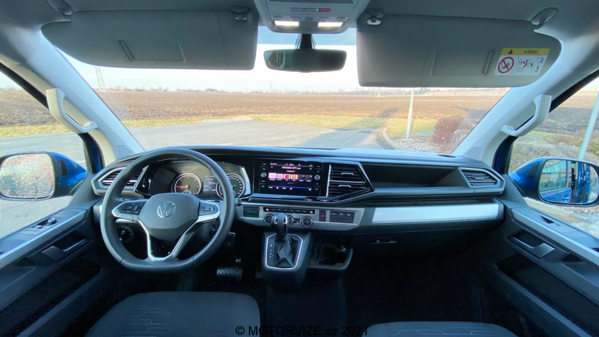 Interior de un VW Nutzfahrzeuge Transporter 2019 (T6.1 Multivan), capturado desde el área del asiento trasero mirando hacia el tablero delantero. La foto está tomada durante el día con luz natural entrando a través del parabrisas delantero, proporcionando una vista detallada del espacio de la cabina, los asientos delanteros, el volante con controles montados, la pantalla táctil central de infoentretenimiento, los botones y perillas del HVAC, el selector de marchas automático, los espacios de almacenamiento, los puertos de conectividad, los controles de puertas y espejos, las características de seguridad y el diseño cómodo de los asientos.