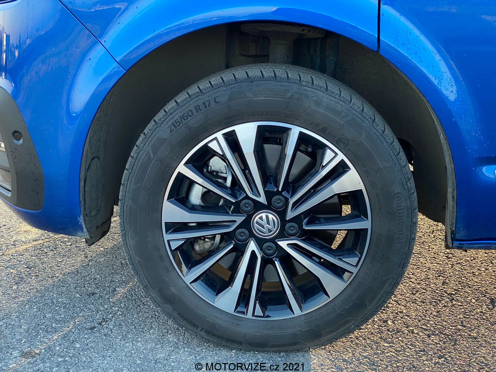 Vista de cerca de una rueda del Volkswagen T6-1 Multivan 2019. Esta imagen de primer plano muestra un diseño de llanta de múltiples radios con acabados alternados en plata y oscuro. La llanta Hankook muestra un patrón de banda de rodadura para rendimiento en calle y especificaciones parciales del neumático. La carrocería azul circundante está en excelente estado con un brillo reflectante. Se observa una altura libre moderada del suelo y pinzas de freno estándar y rotores parcialmente visibles. La luz natural resalta el enfoque en la llanta, mientras que el fondo está desenfocado debido a la toma en ángulo bajo.