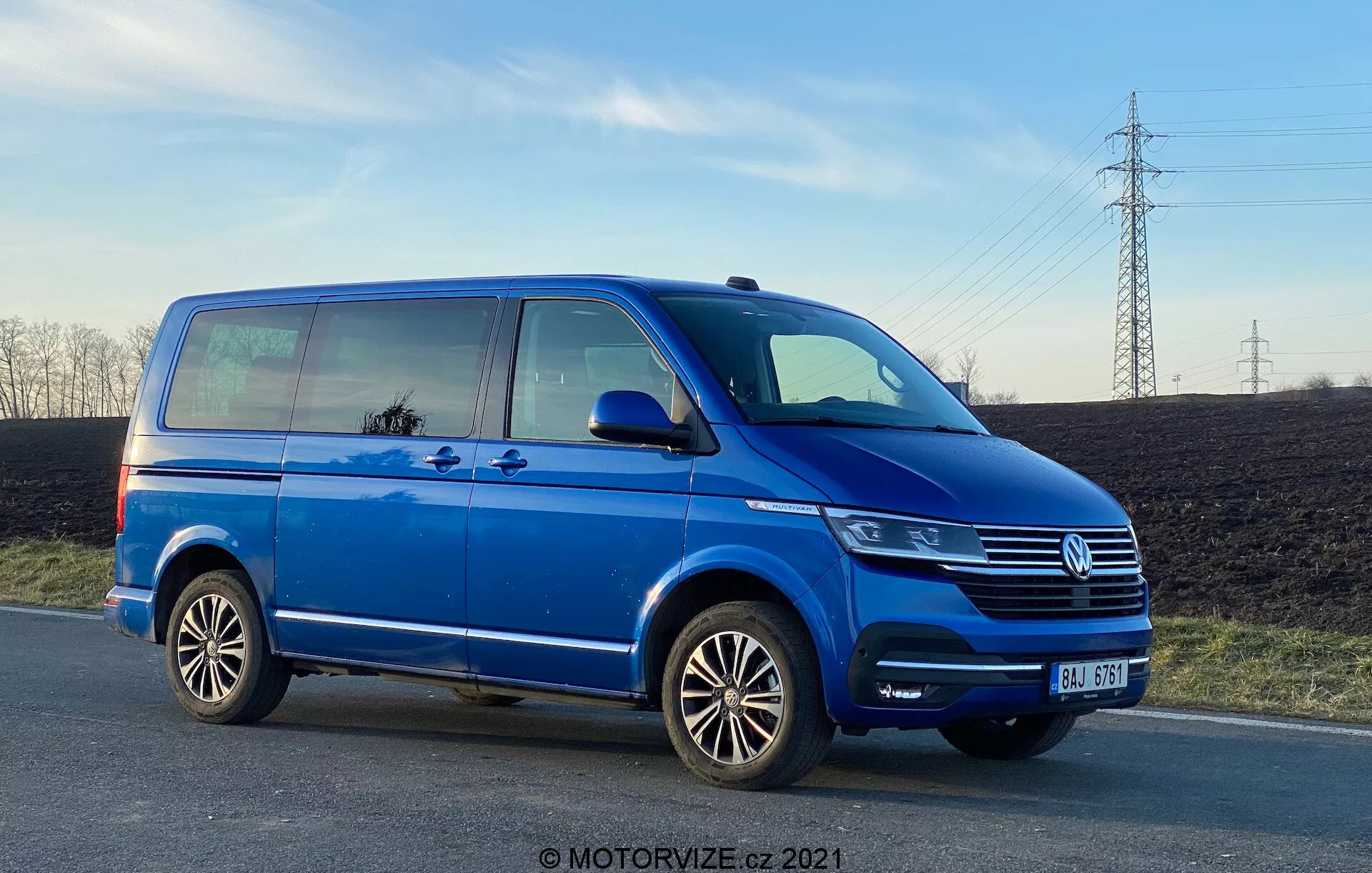 Una furgoneta Volkswagen Transporter T6 azul, mostrando su perfil lateral desde un ángulo bajo, resaltando su longitud y altura. La imagen presenta detalles como la puerta corrediza del pasajero, la puerta delantera del pasajero, la rueda trasera del lado del pasajero, llantas de aleación multirradio y vistas parciales del faro, la parrilla Volkswagen con el emblema y el parachoques delantero. La foto, tomada a última hora de la tarde, muestra la distancia al suelo y refleja el cielo y los alrededores en las ventanas, con un cielo claro y cables de electricidad en el fondo.