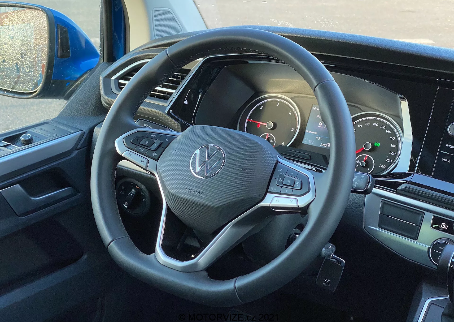 Vista interior del VW Nutzfahrzeuge Transporter 2019 (T6-1 Multivan) desde la perspectiva del conductor, enfocándose en el volante y el salpicadero. El volante muestra el emblema de Volkswagen y está equipado con revestimiento de cuero, botones multifuncionales y palancas de cambio. El cuadro de instrumentos parcialmente visible incluye diales analógicos y una pantalla digital. La pantalla central de infoentretenimiento y los controles de climatización son ligeramente visibles, indicando características modernas de multimedia y confort.